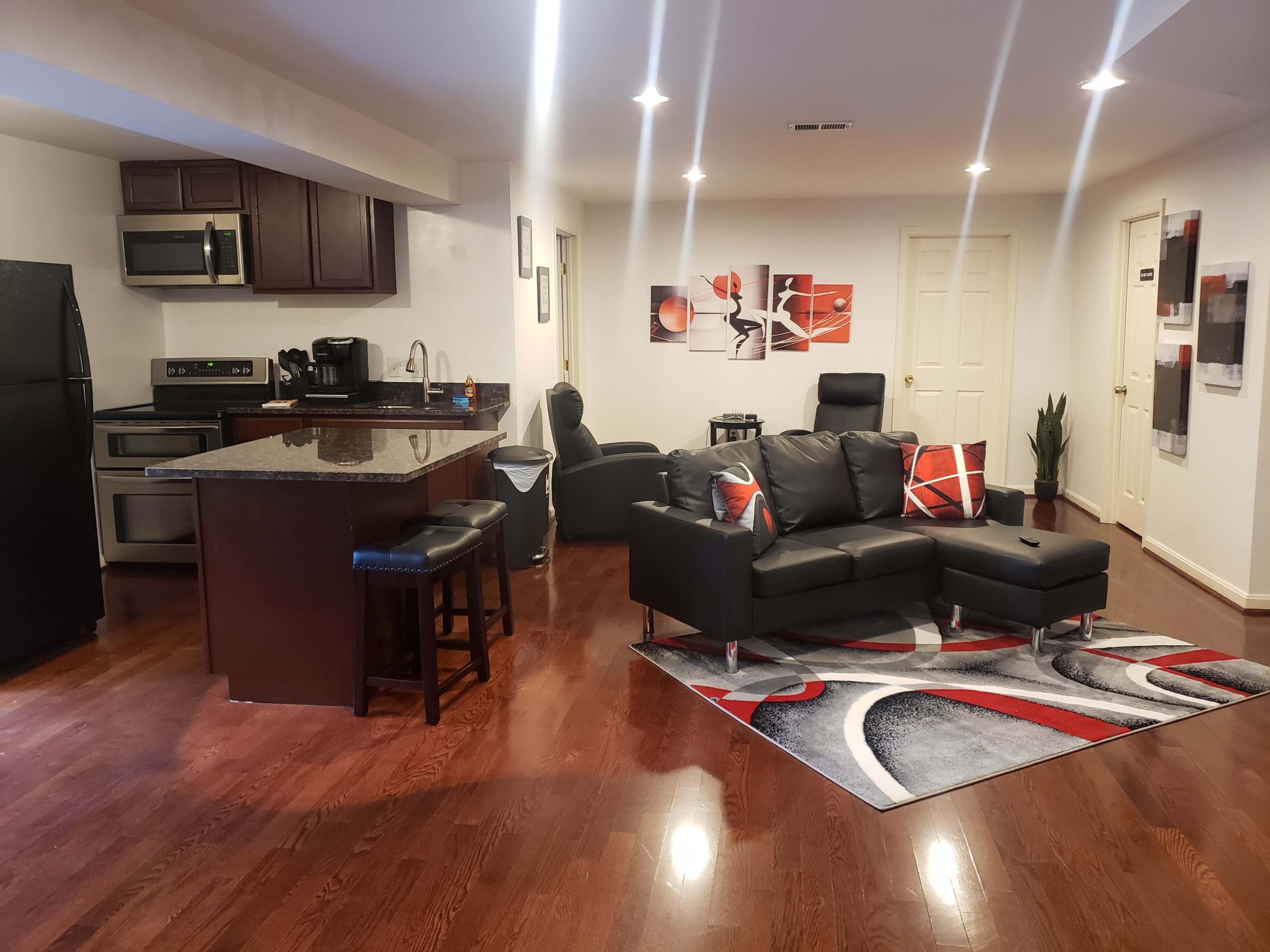 A modern open-concept living space featuring a kitchen with dark wood cabinets, stainless steel appliances, and a living area with a black sectional sofa and decorative rug.