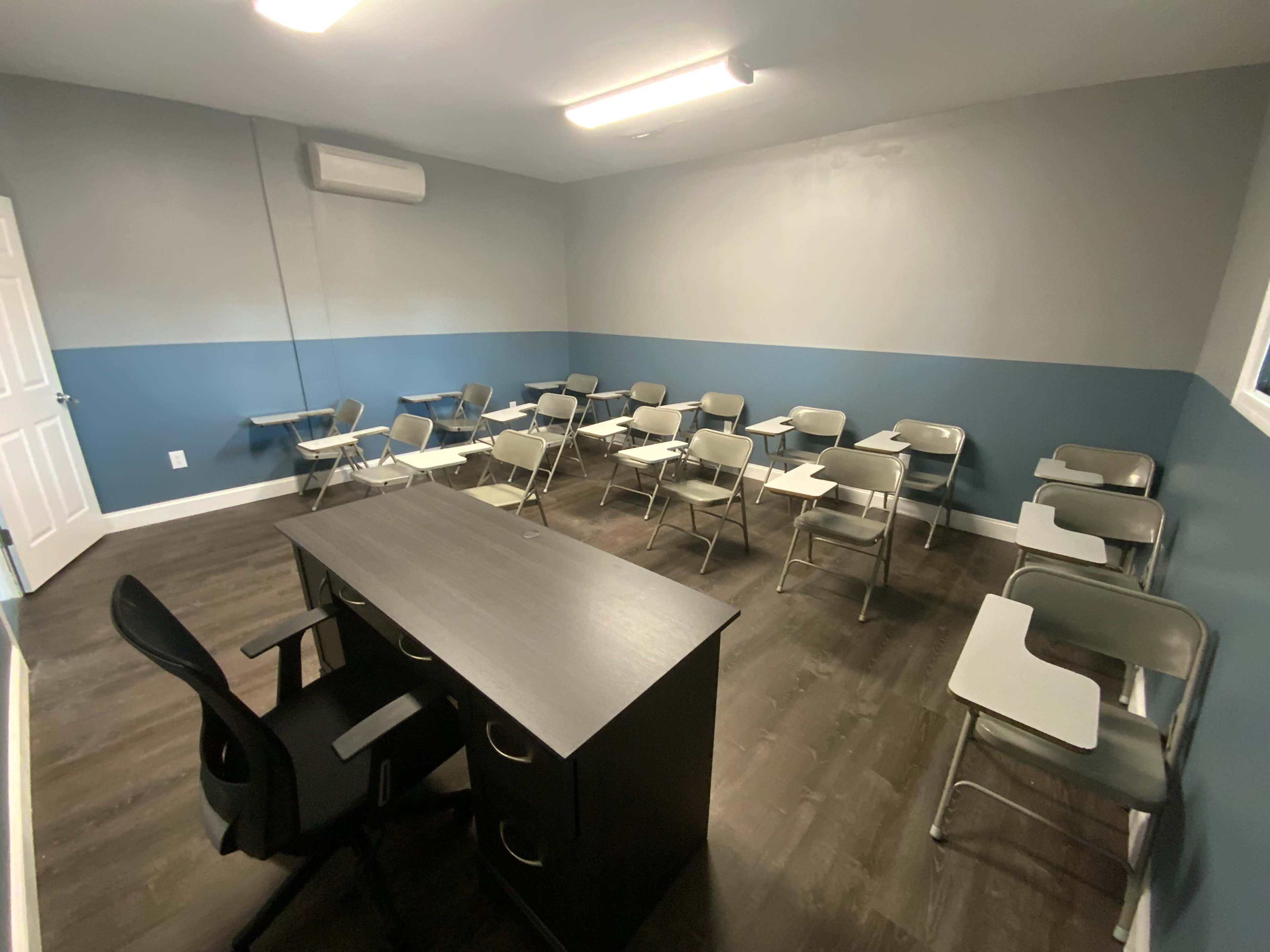A classroom with gray walls and a blue accent, featuring several metal desks arranged facing a front desk.