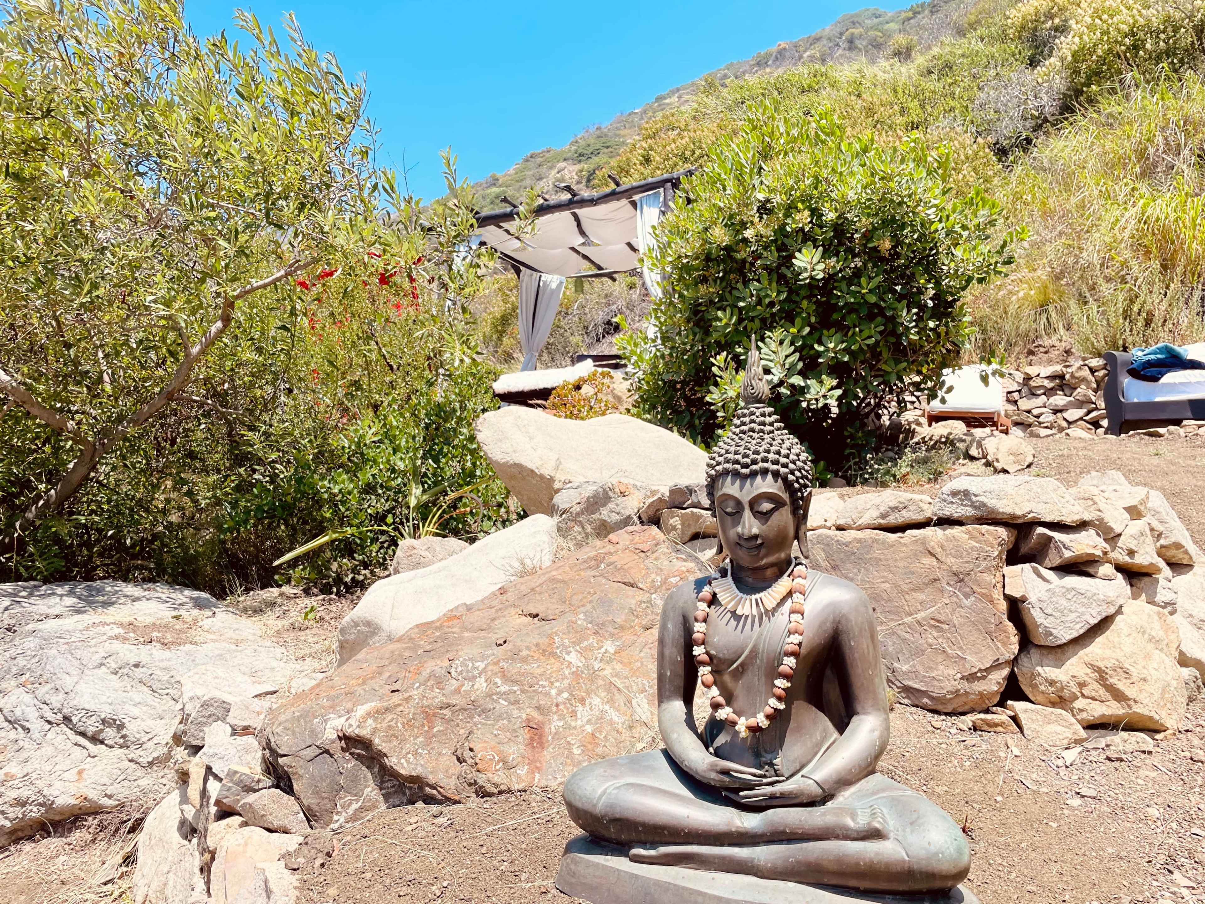 A Buddha statue sits on a rock in a garden surrounded by greenery and a glimpse of a shaded seating area in the background.