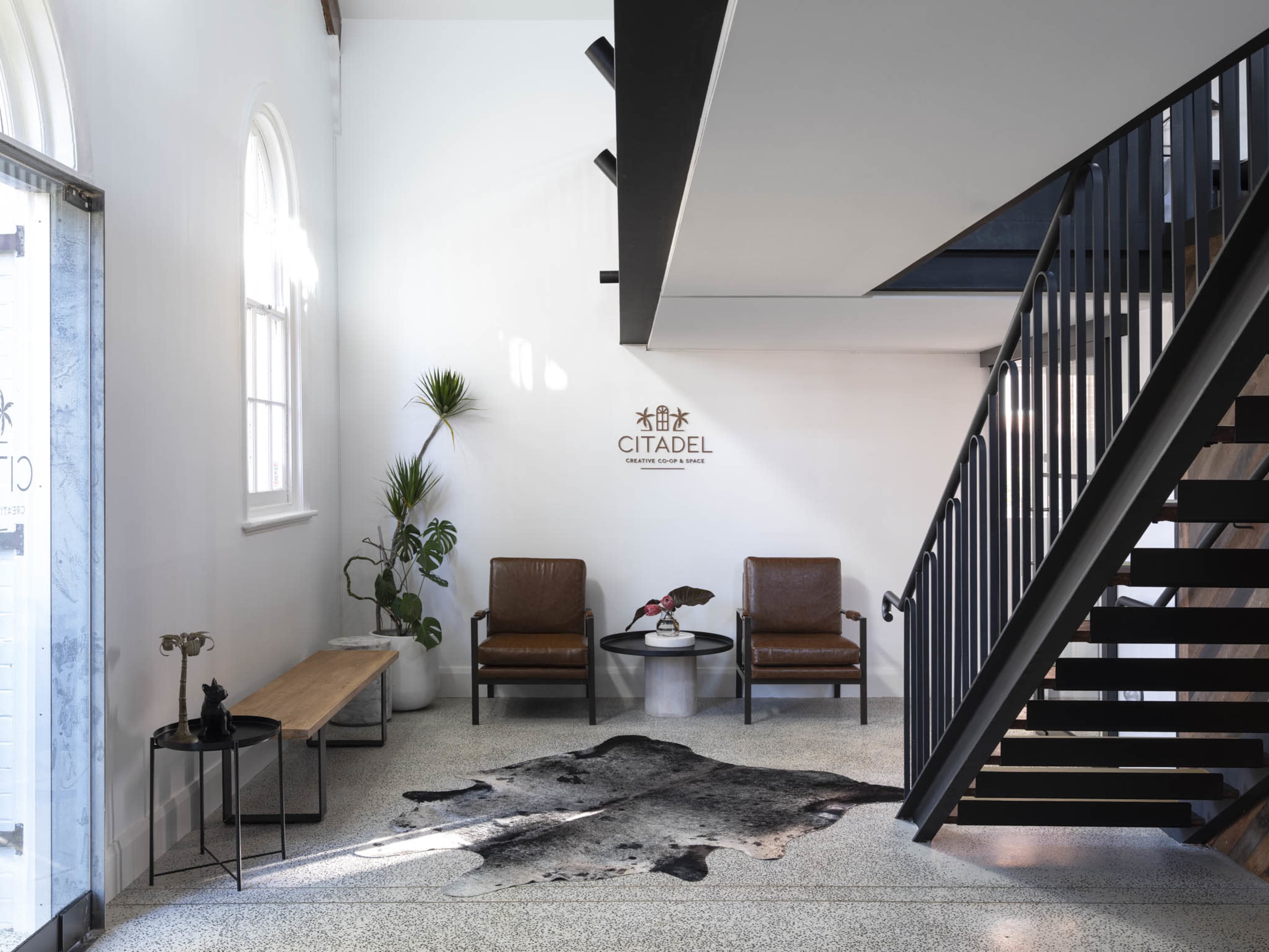 The image shows a modern lobby area with two leather chairs, a small side table, a wooden bench, a potted plant, and a staircase adjacent to a wall that features the word "CITADEL."