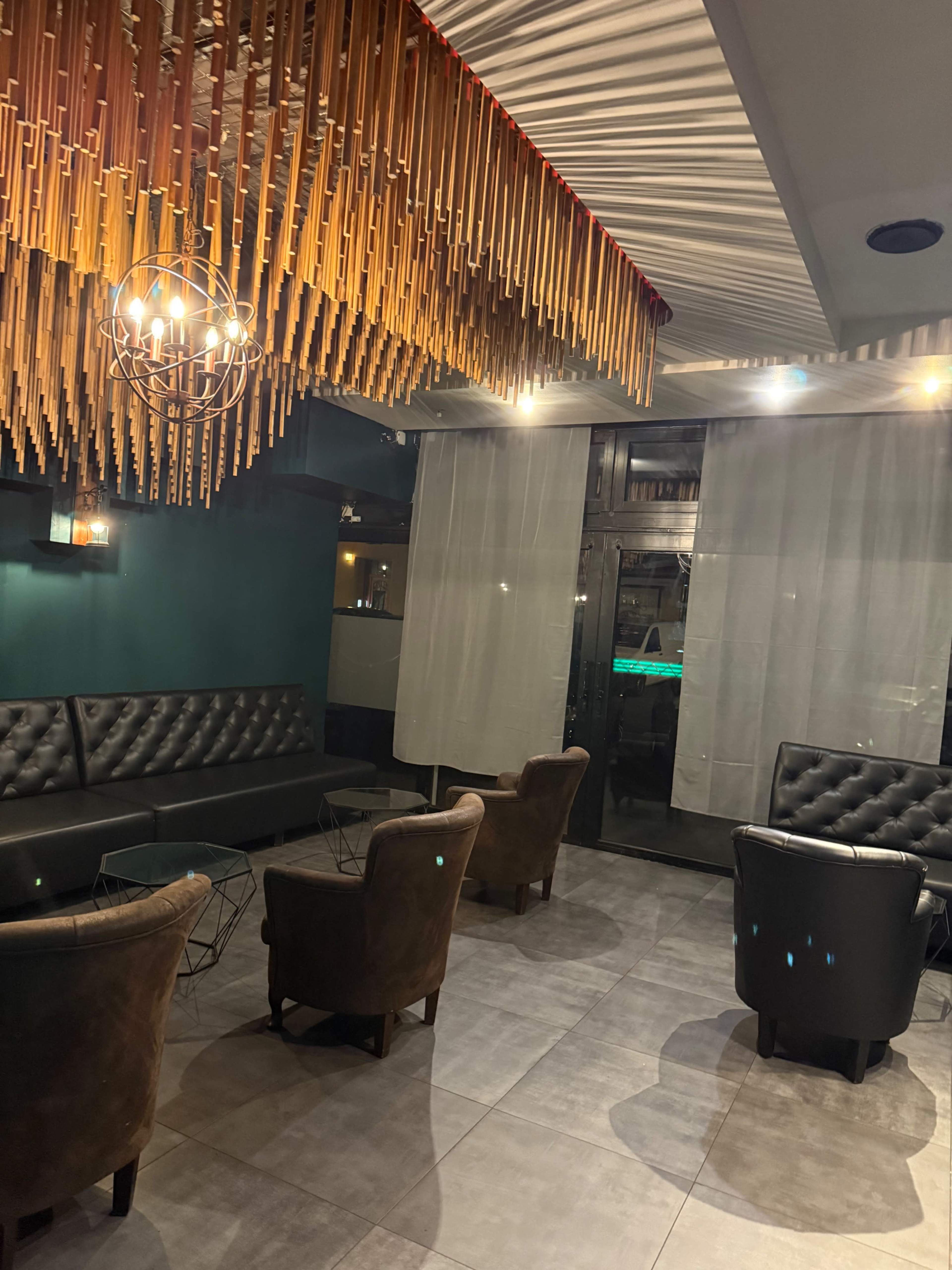 The image shows a modern lounge area with dark leather seating, a decorative overhead light fixture, and large windows covered by sheer curtains.