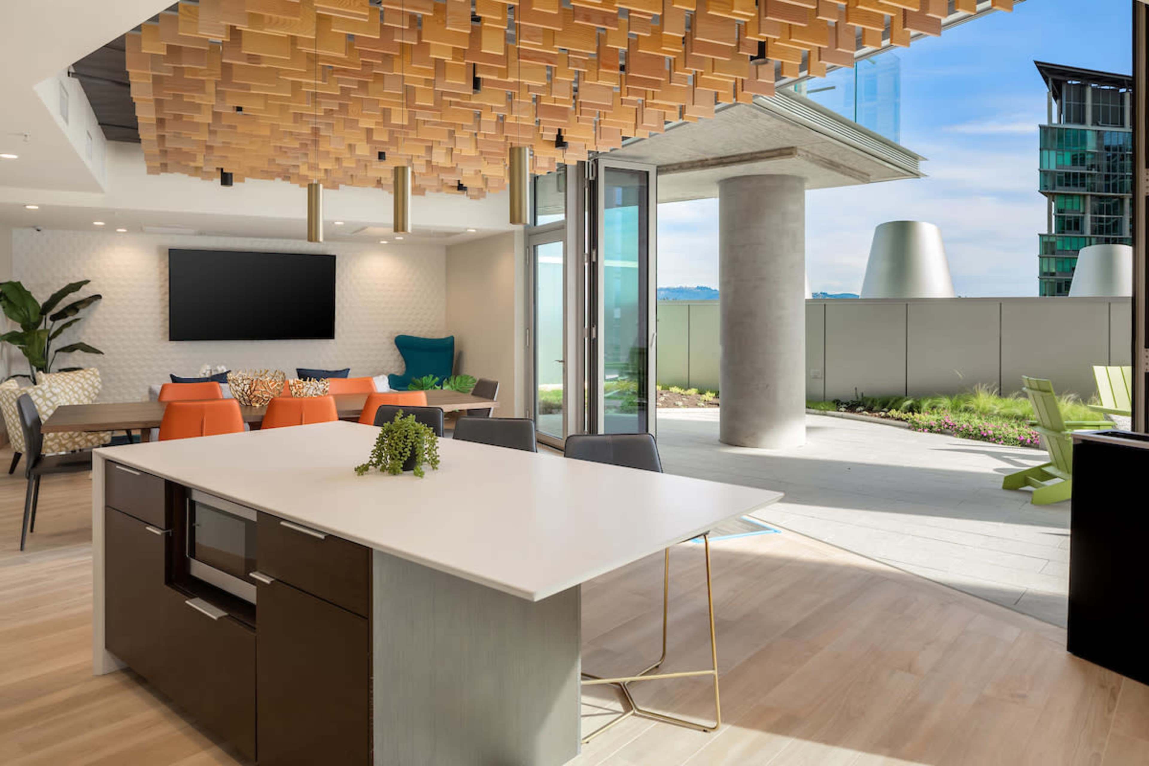 The image shows a modern open-concept lounge area with a large island table, stylish seating, and sliding glass doors leading to a patio with greenery.