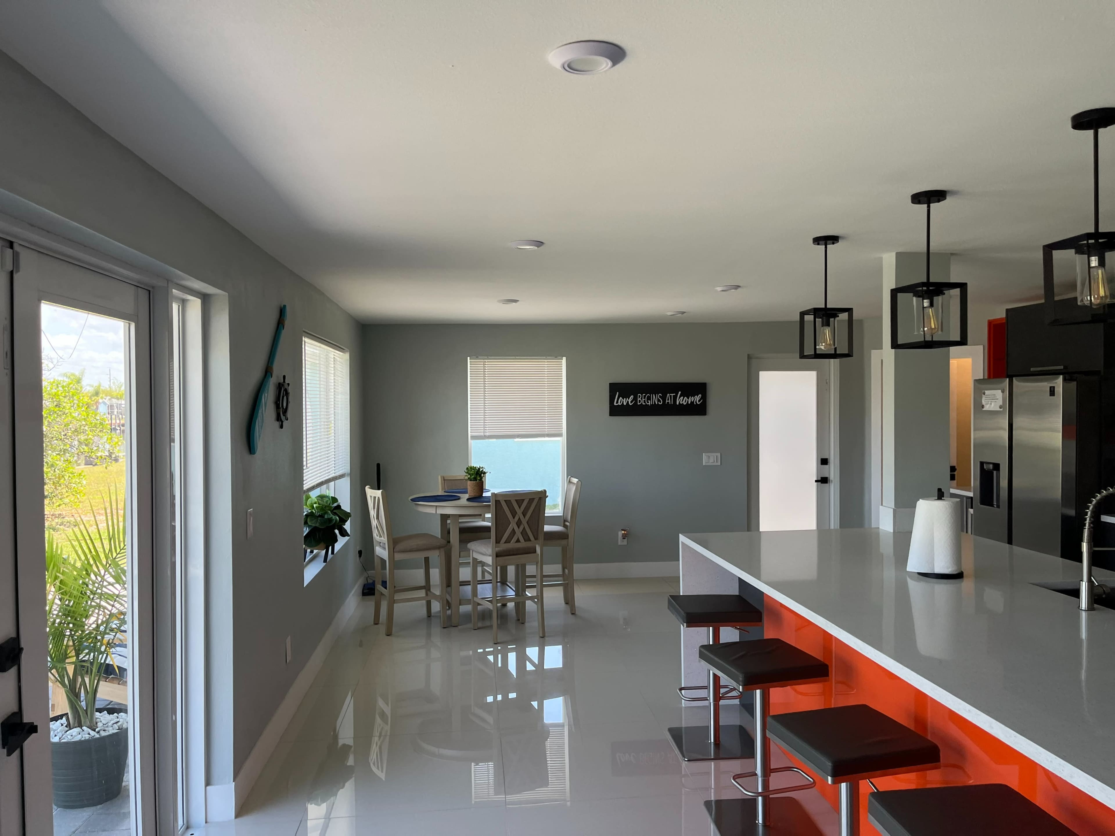 The image shows a modern kitchen and dining area with a large island, sleek bar stools, and large windows allowing natural light in.