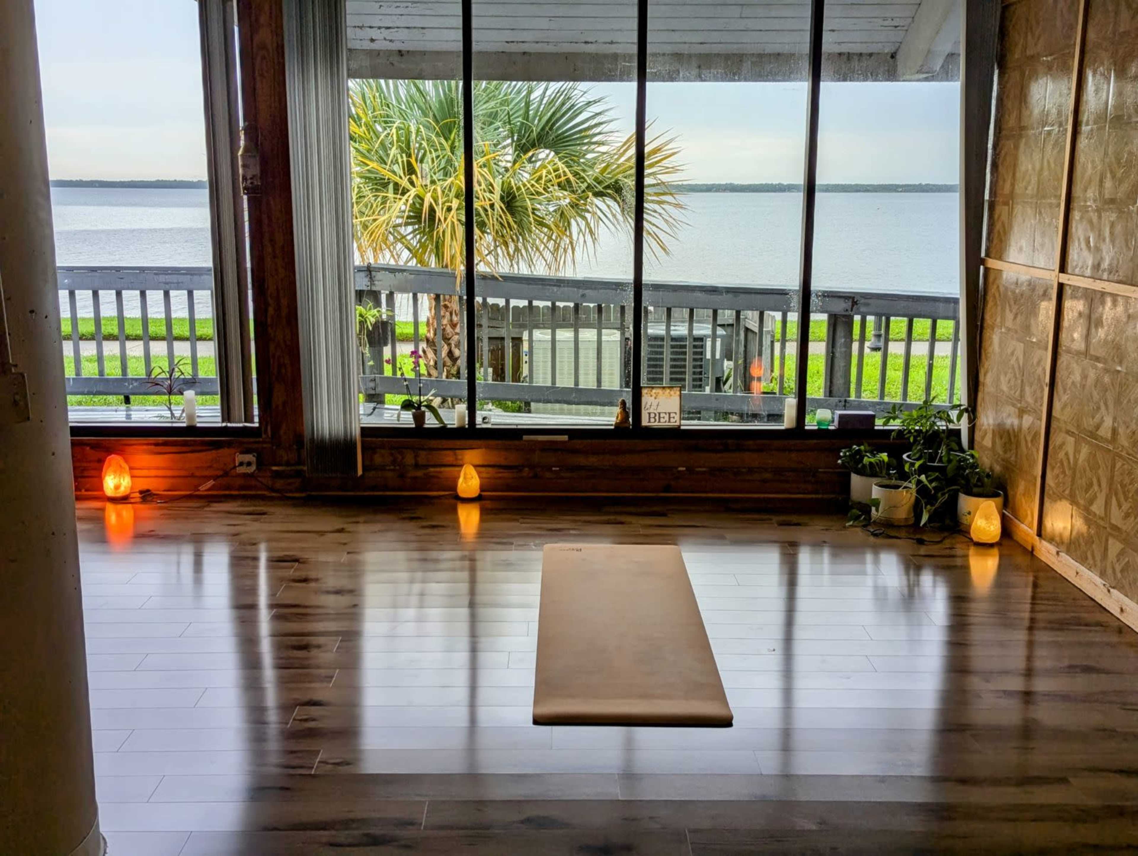 A yoga mat is placed on a wooden floor in a serene studio that features large windows overlooking a body of water and palm trees.