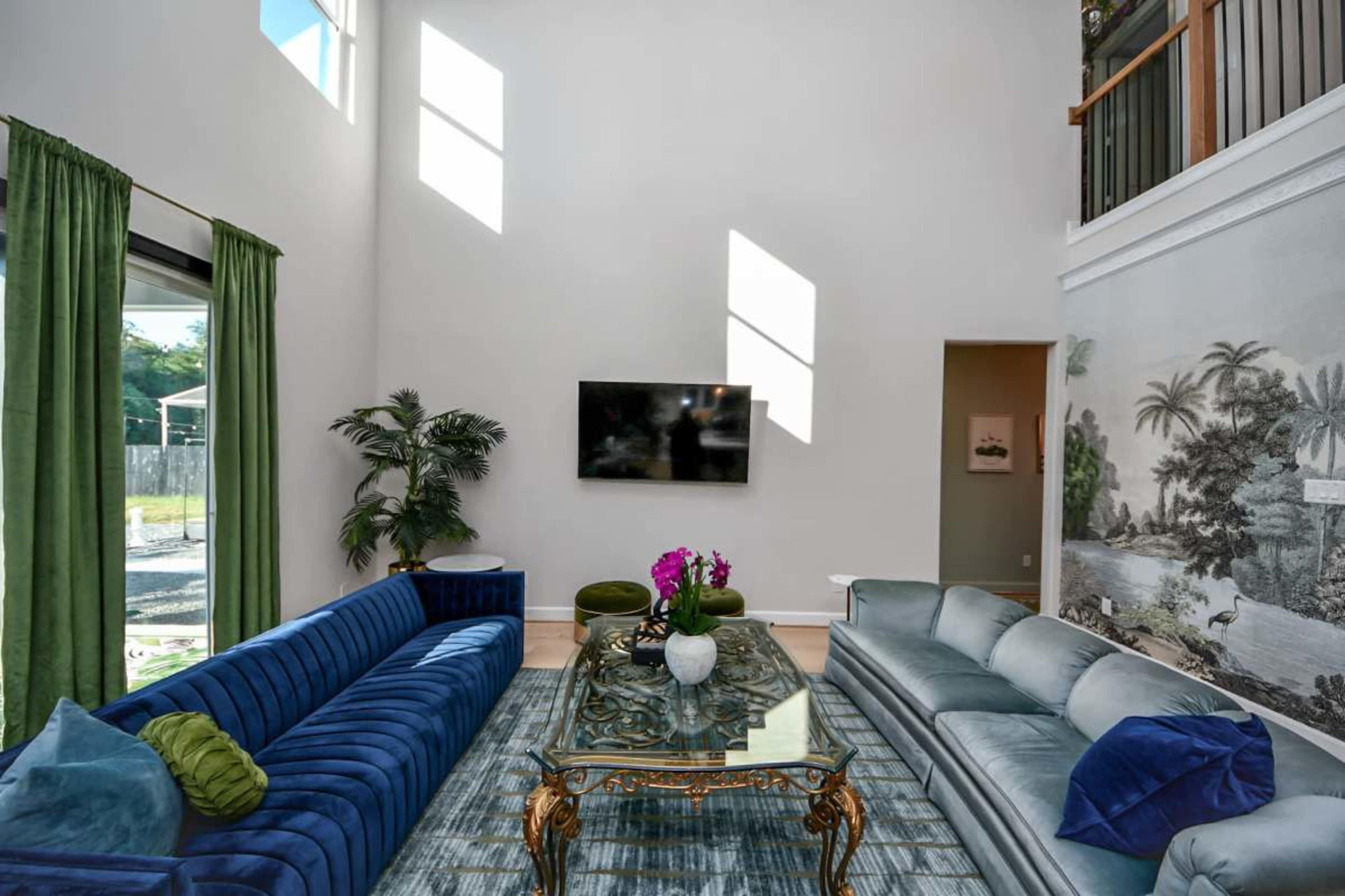 A spacious living room features a tall ceiling, large windows letting in natural light, a blue velvet sofa, a light gray sofa, and a decorative glass coffee table with floral arrangements.
