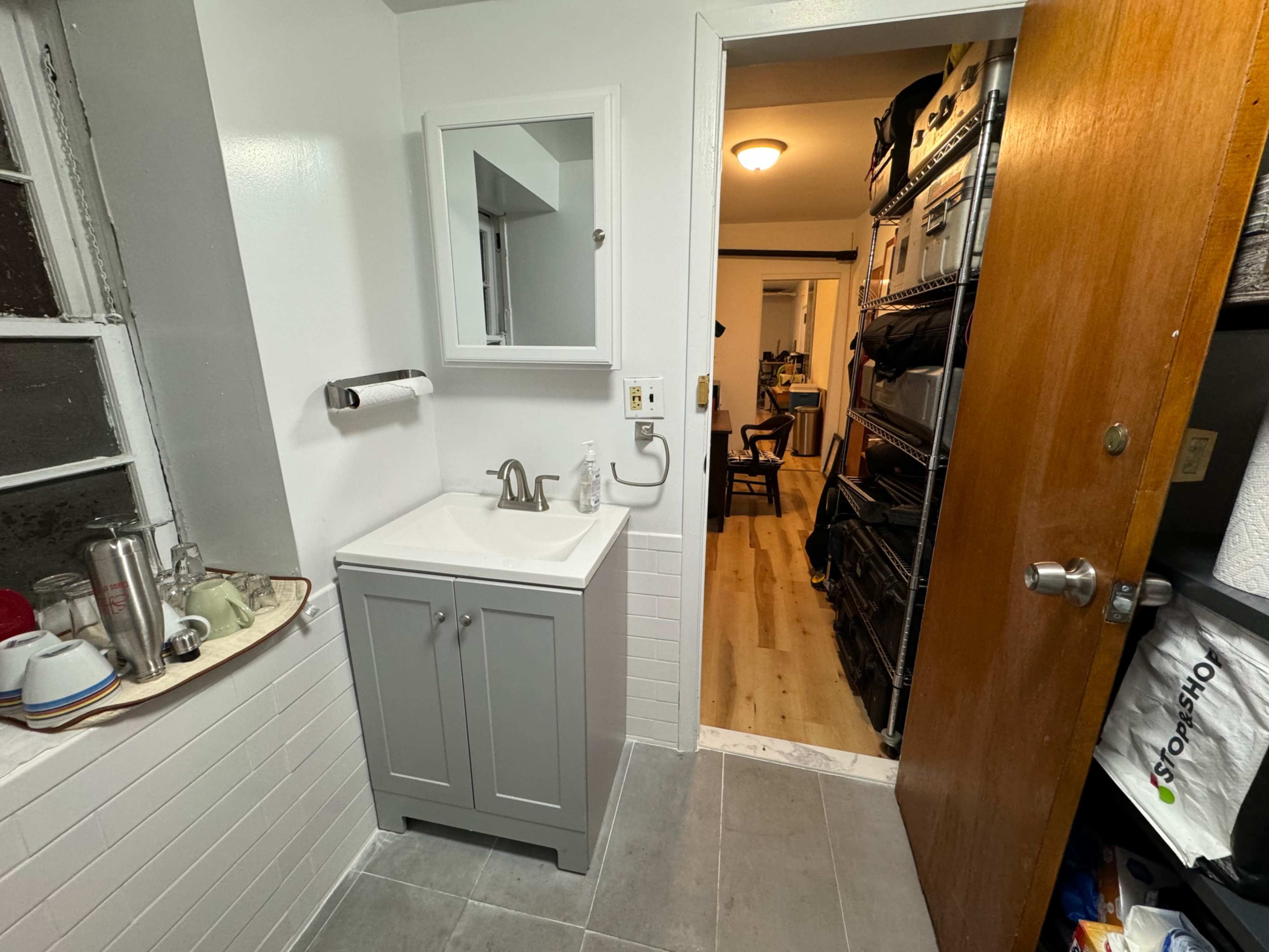 The image shows a small bathroom with a gray sink cabinet and a doorway leading to a storage area.
