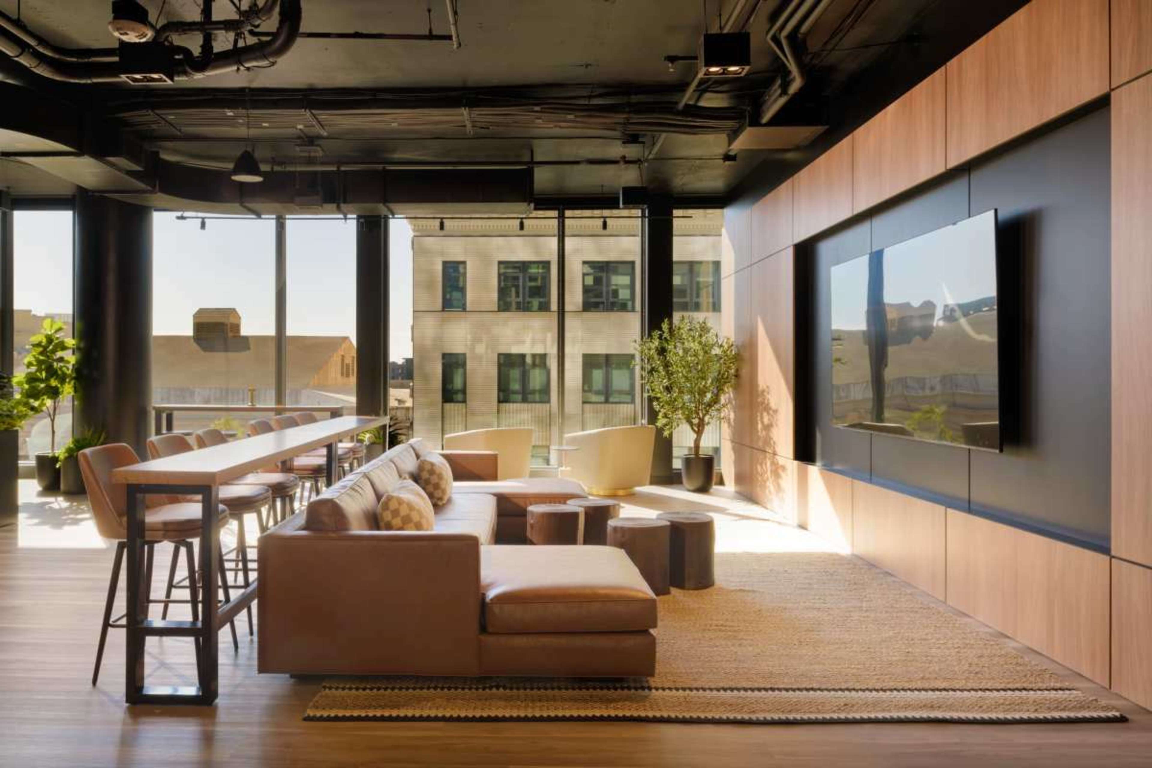 The image shows a modern lounge area with a sectional sofa, bar-style seating, and a large television mounted on a wooden wall.