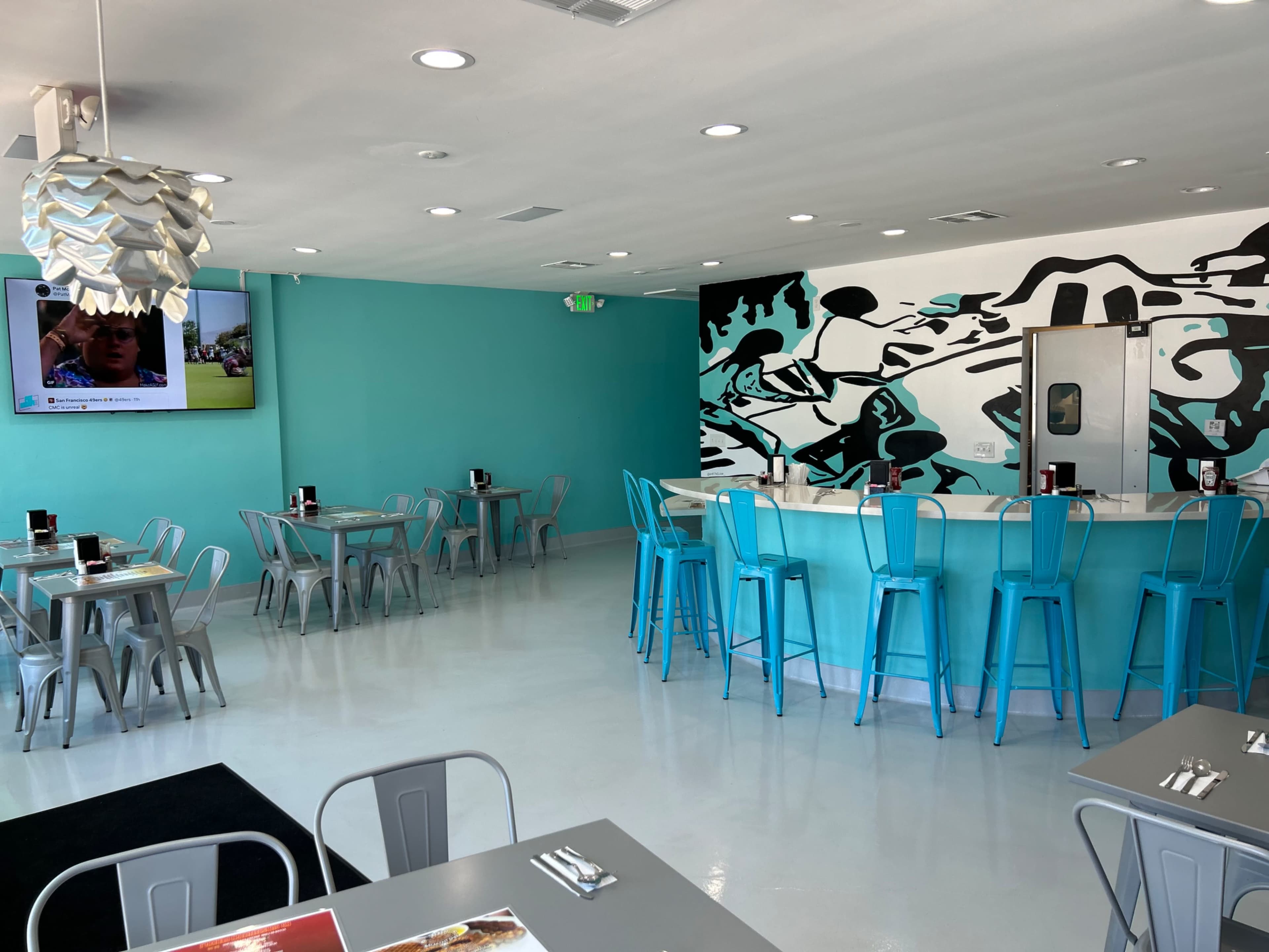 The image shows a modern restaurant interior with teal walls, a bar area, and several tables with metal chairs.