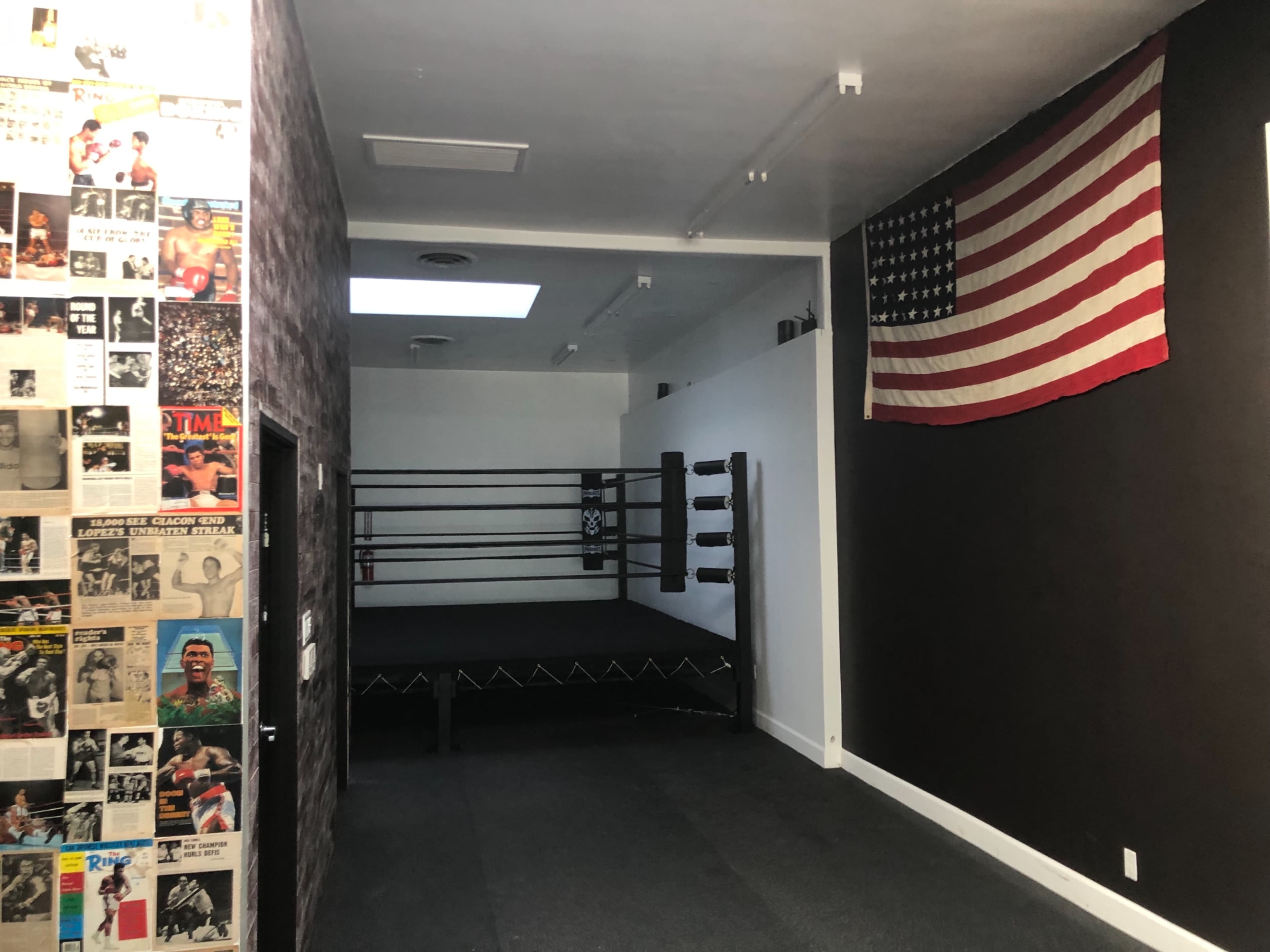 A boxing gym featuring a boxing ring, an American flag on the wall, and a collage of old boxing posters on the adjacent wall.