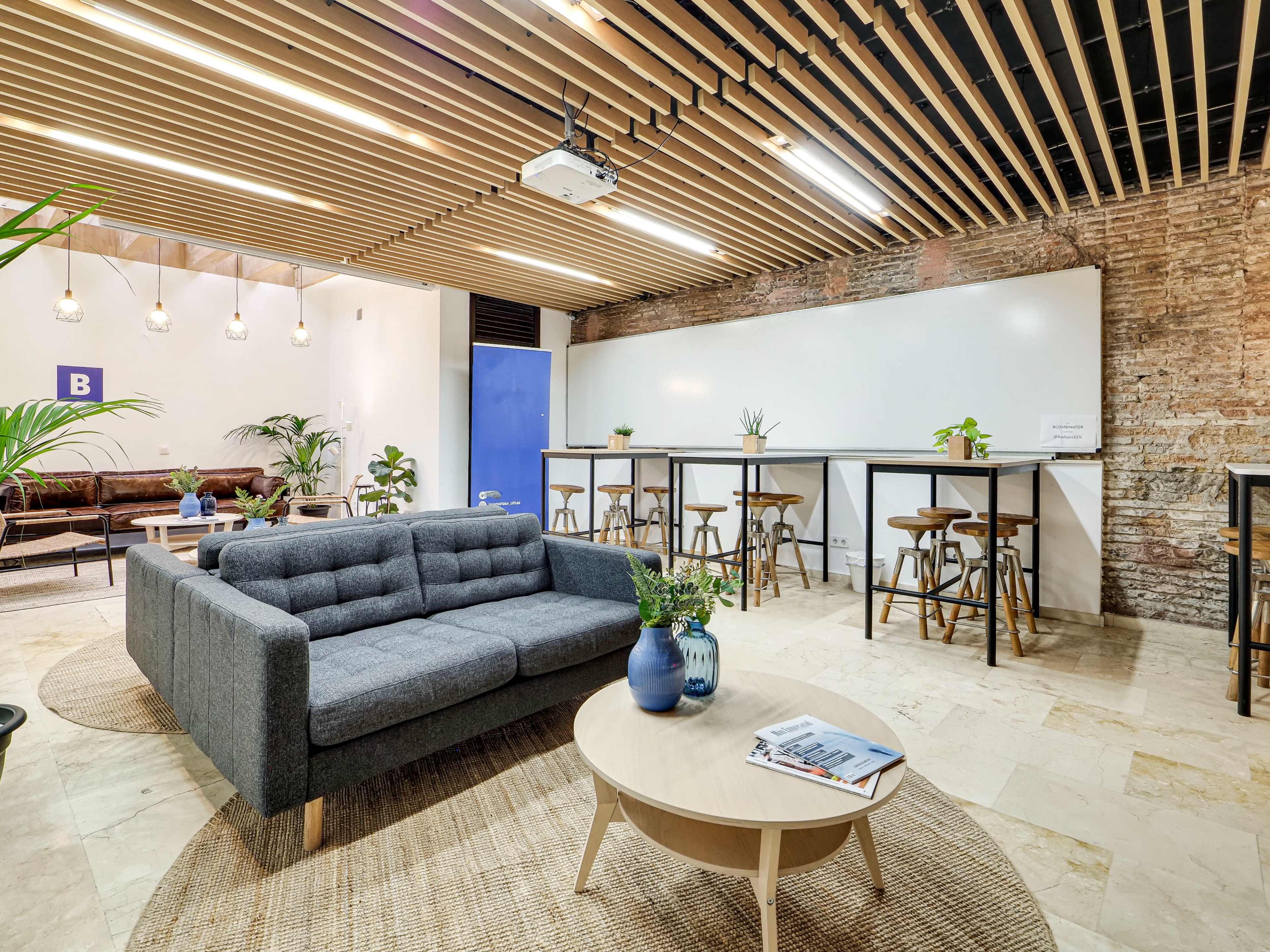 The image shows a modern lounge area with a gray sofa, a round coffee table, and high stools arranged around a bar-style counter, featuring wooden beams on the ceiling and plants throughout.