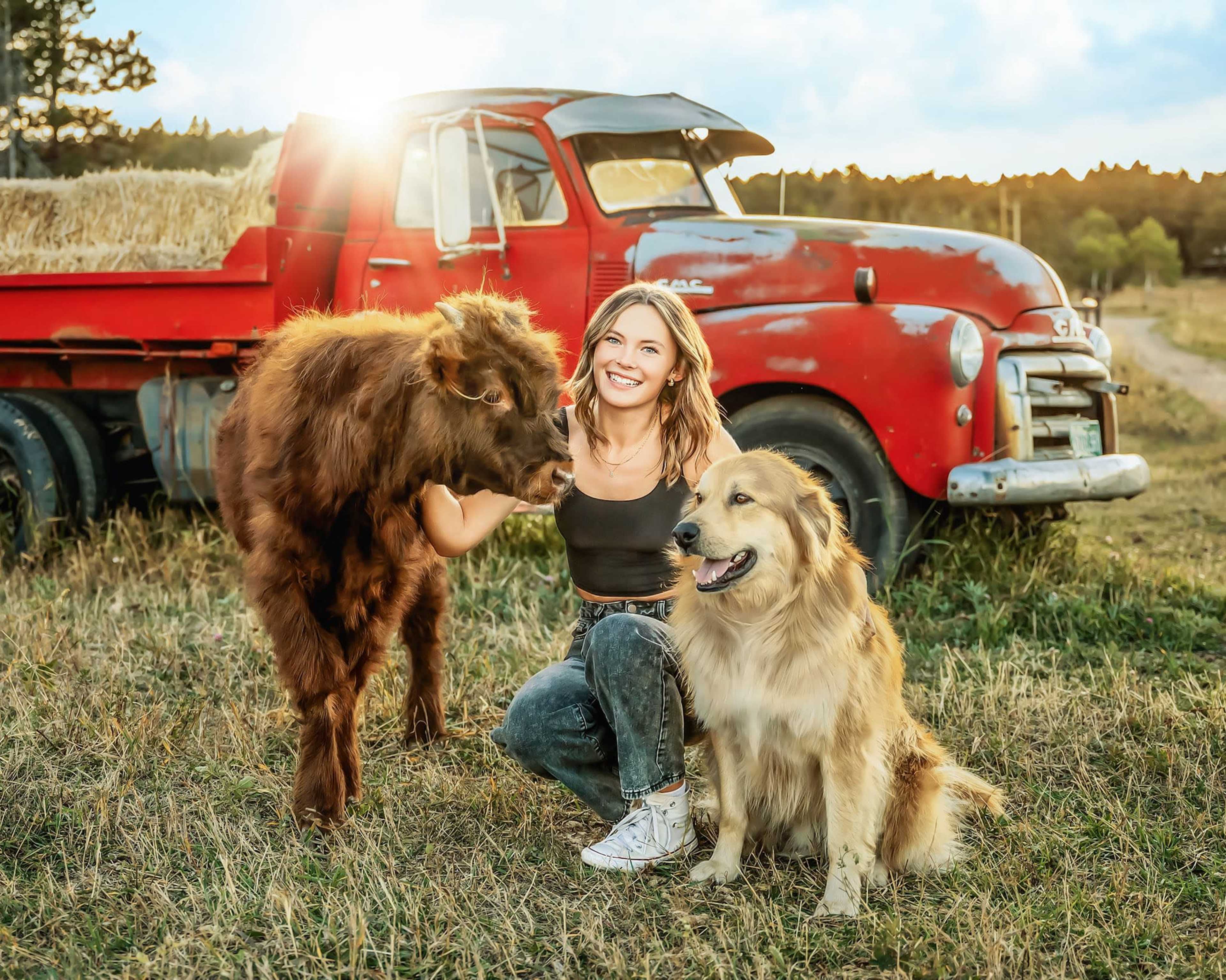 A woman crouches in a field next to a calf and a dog, with a vintage red truck in the background.