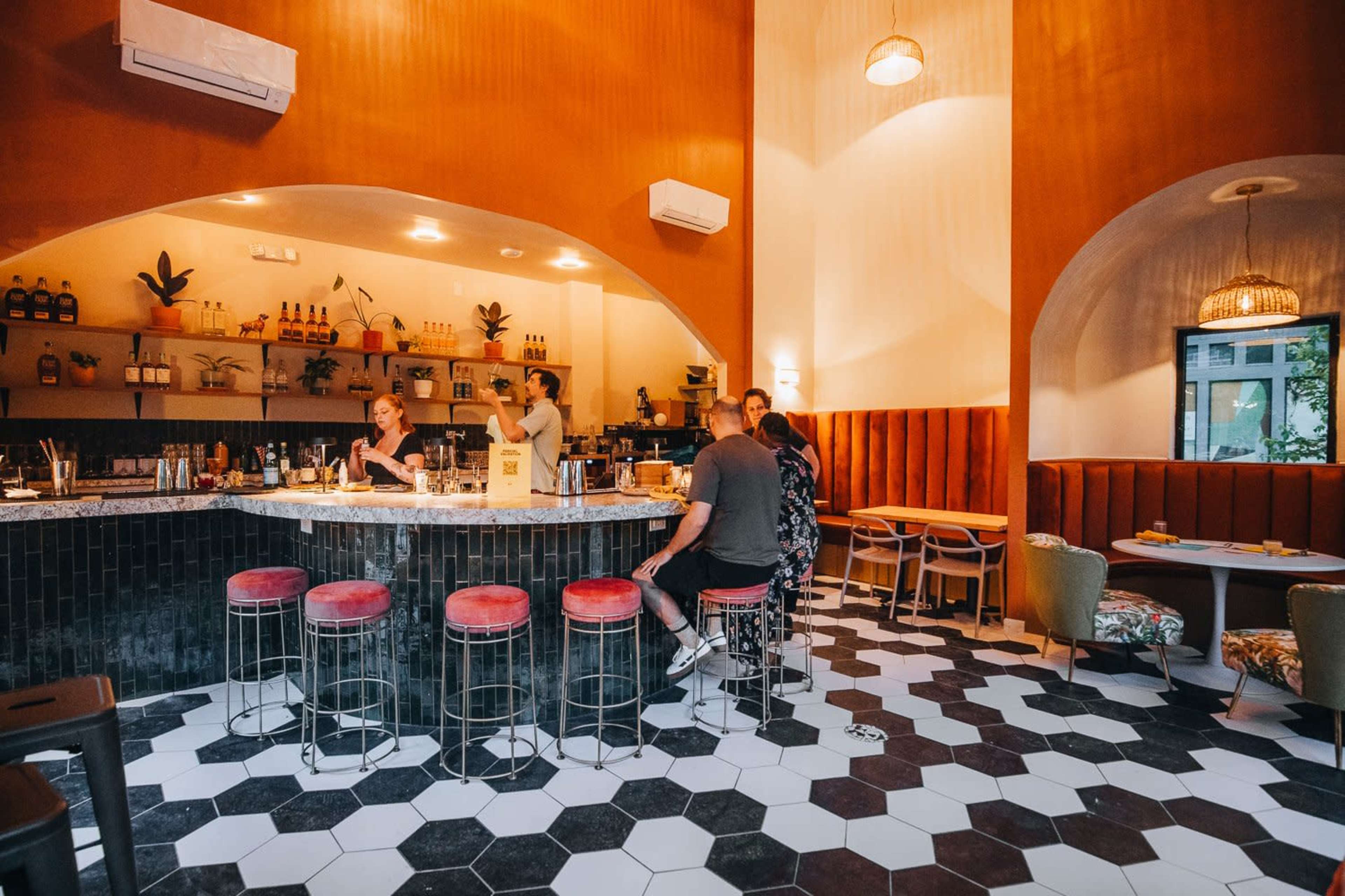 The image shows a stylish bar area with patrons seated at a hexagonal-patterned floor, surrounded by shelves of drinks and plants.
