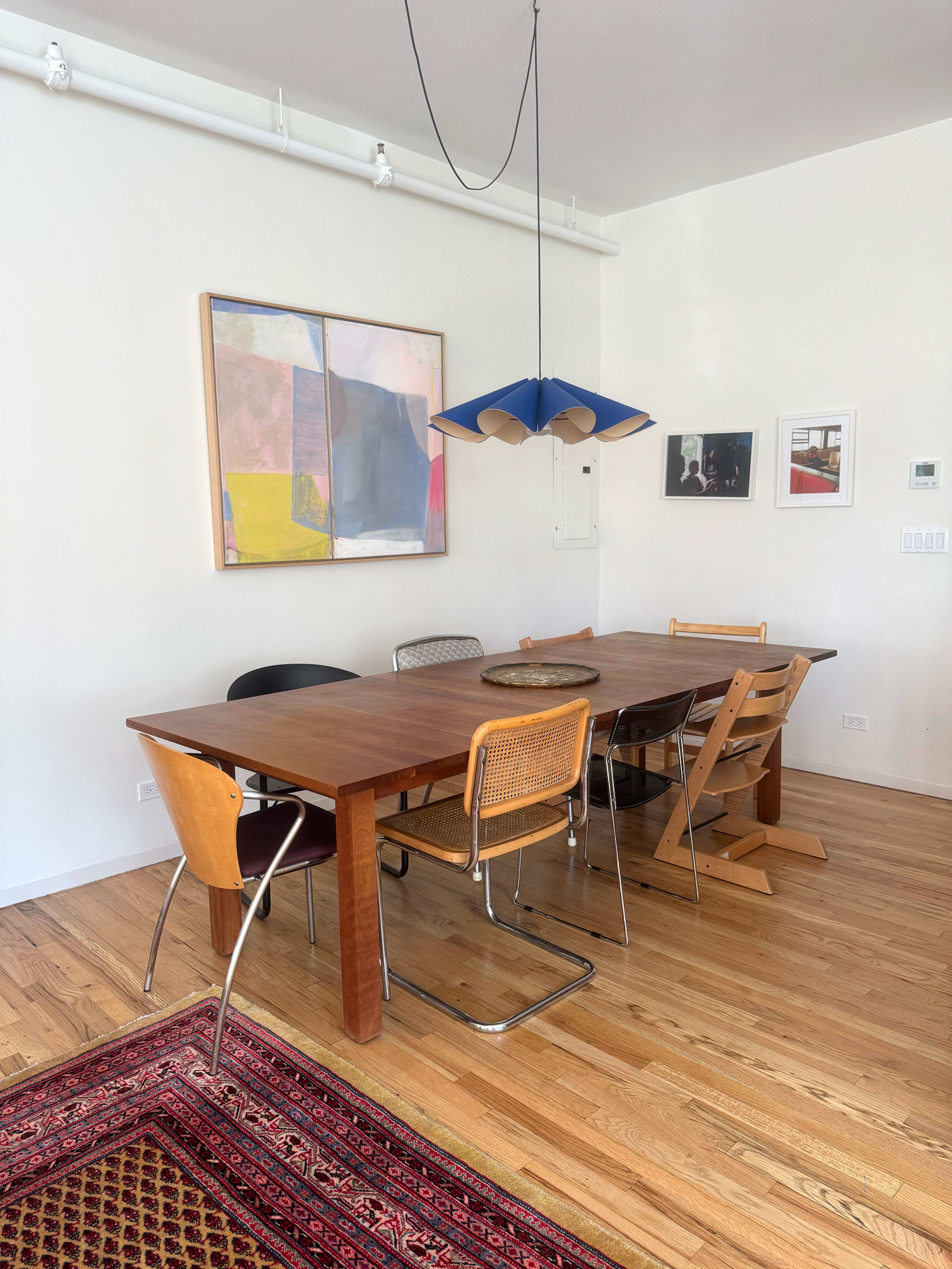 A wooden dining table with mixed chairs is set in a brightly lit room featuring abstract artwork on the walls and a patterned rug on the floor.