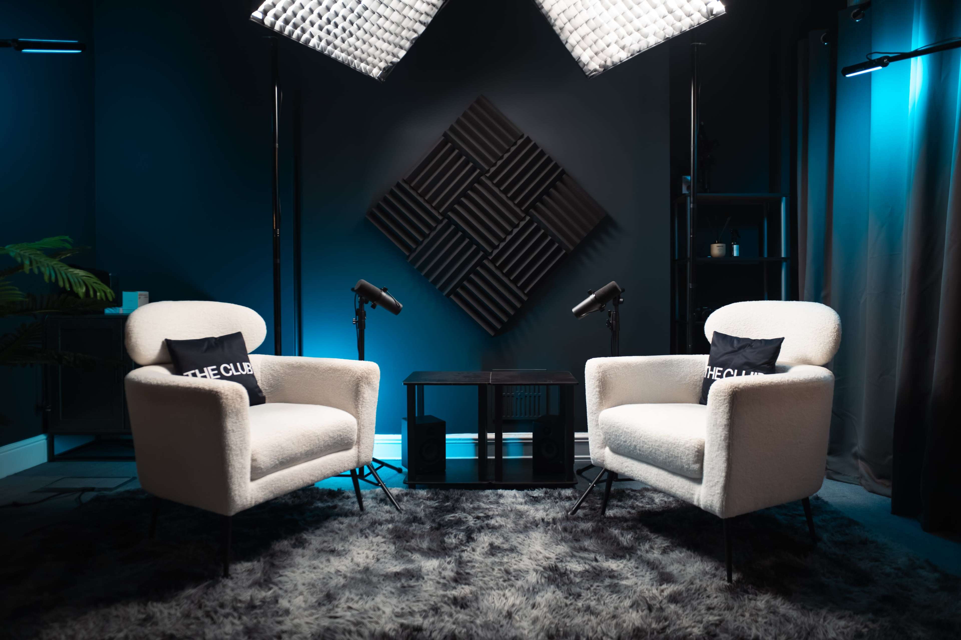 The image shows two white chairs positioned on a plush gray rug under bright lighting, with sound-absorbing panels on the wall behind them.