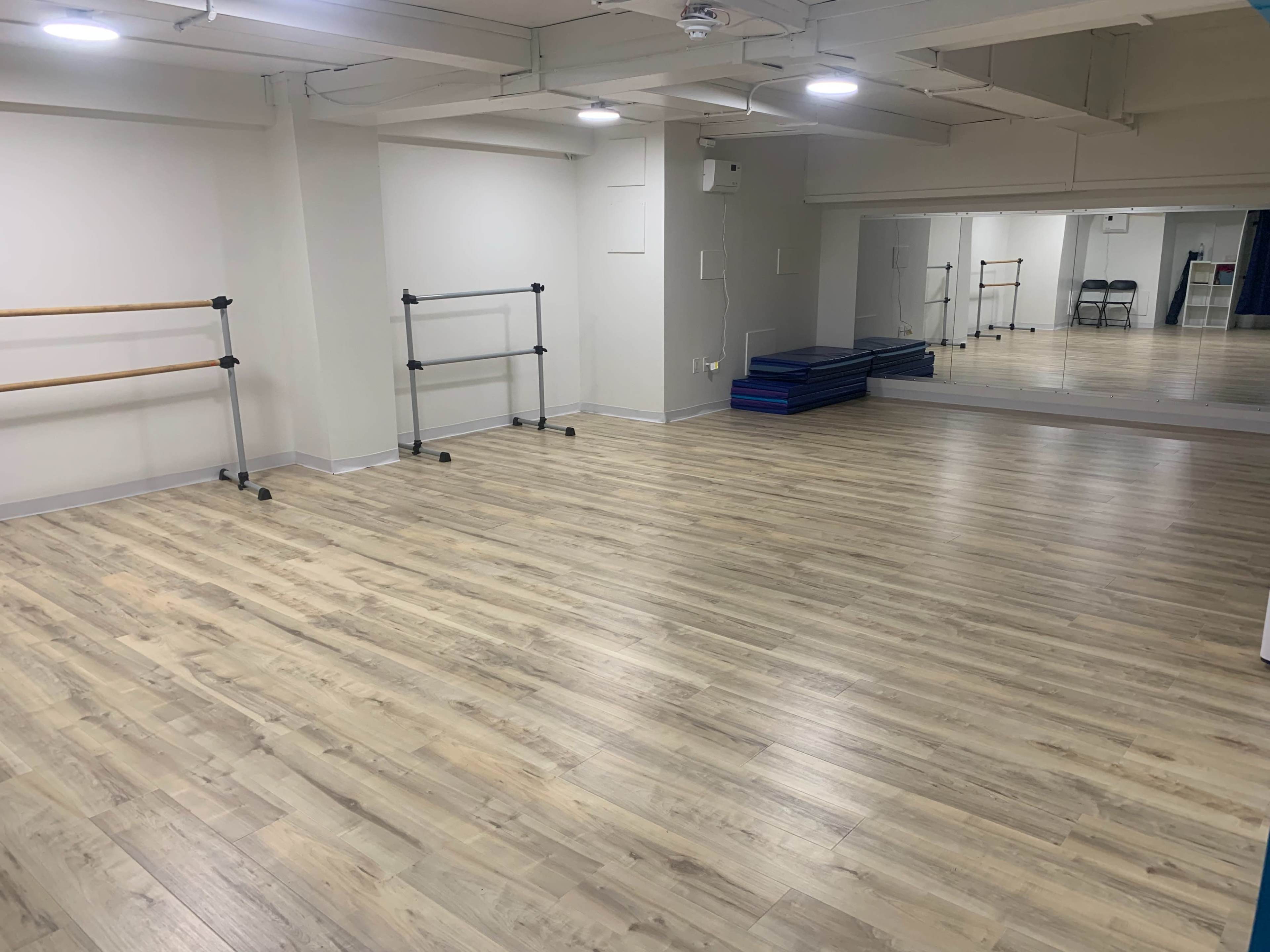 The image shows a spacious, brightly lit exercise studio with wooden flooring, barre equipment against the walls, and a large mirror on one side.