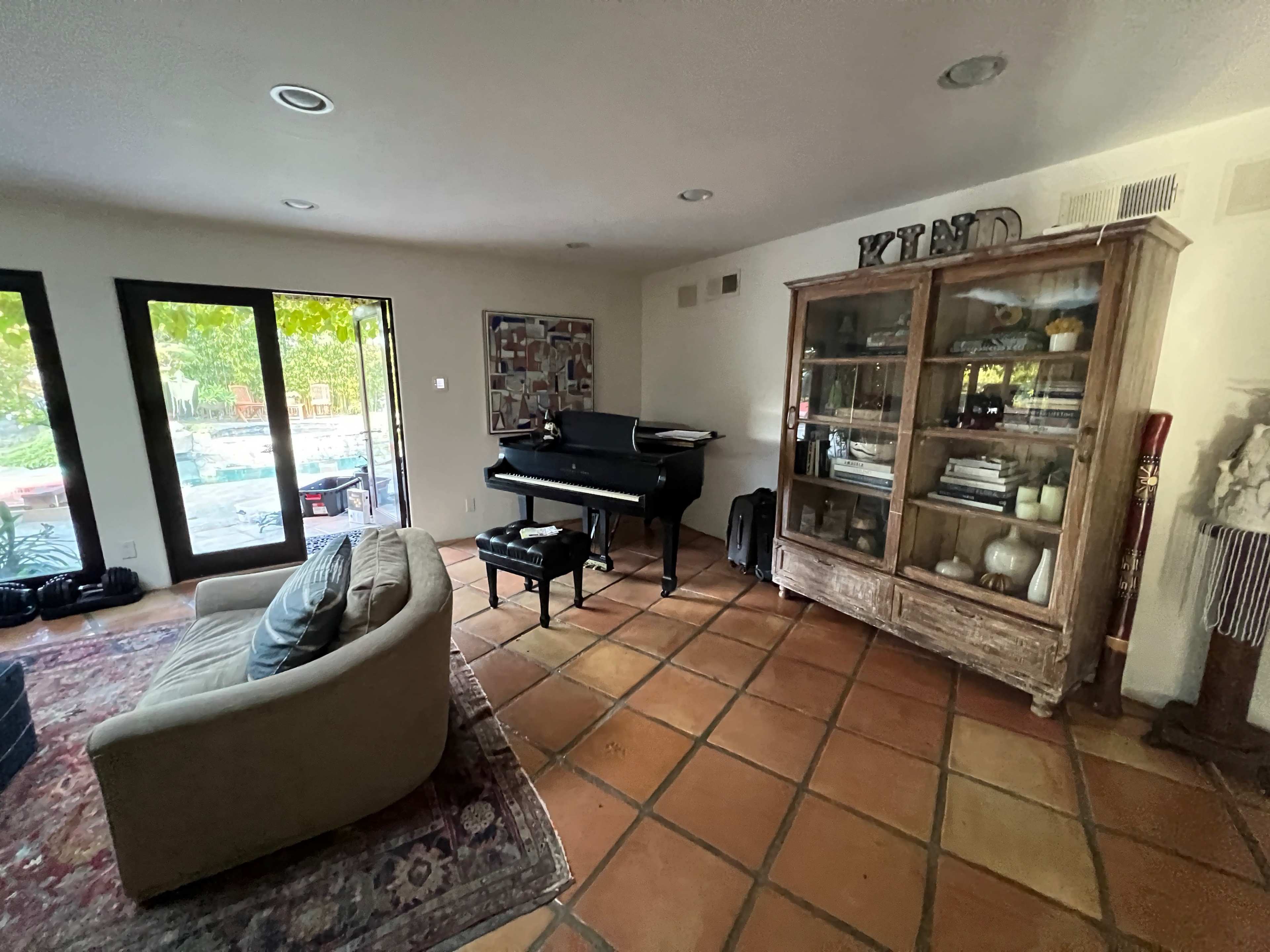 The image shows a living room with a piano, a glass-fronted cabinet, and a sofa, featuring terracotta tile flooring and large windows overlooking a pool area.