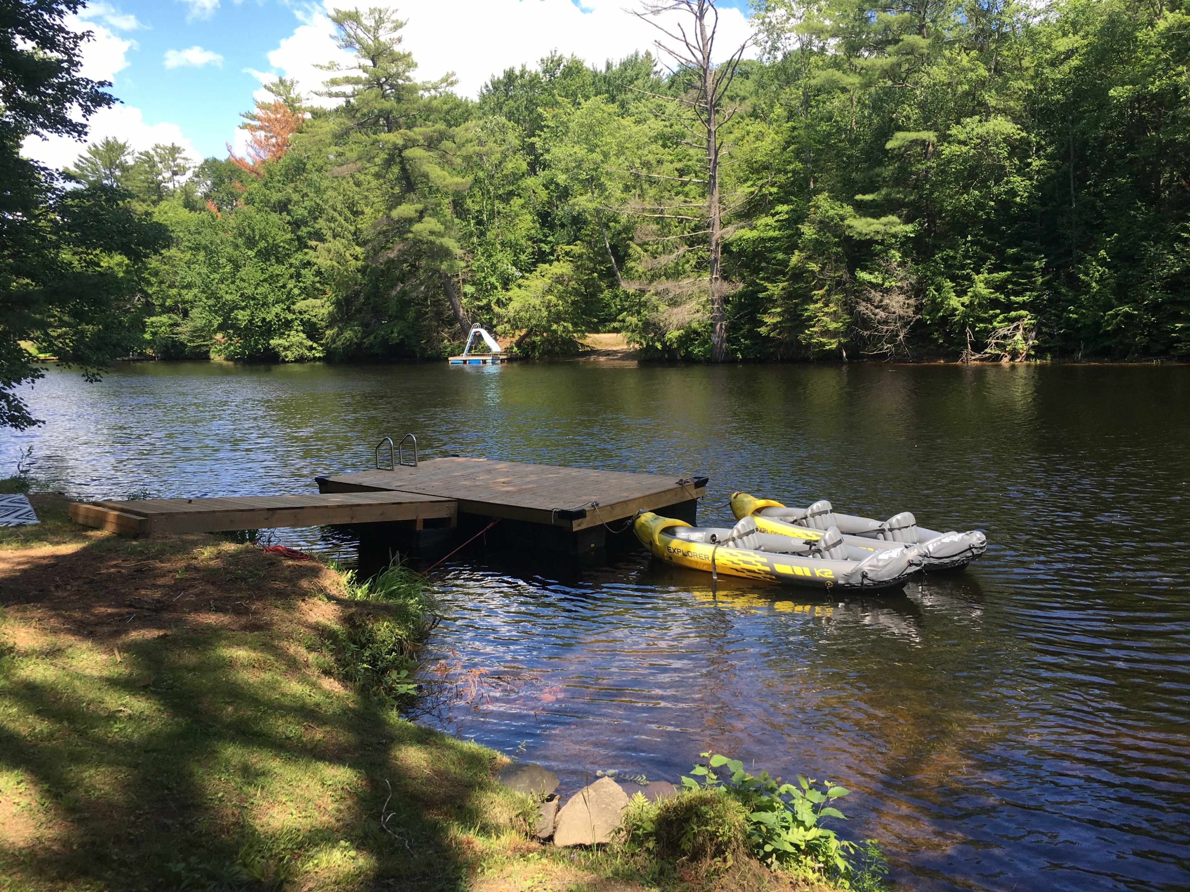 A wooden dock extends into a calm river, with several inflatable rafts tied to it and lush greenery surrounding the area.