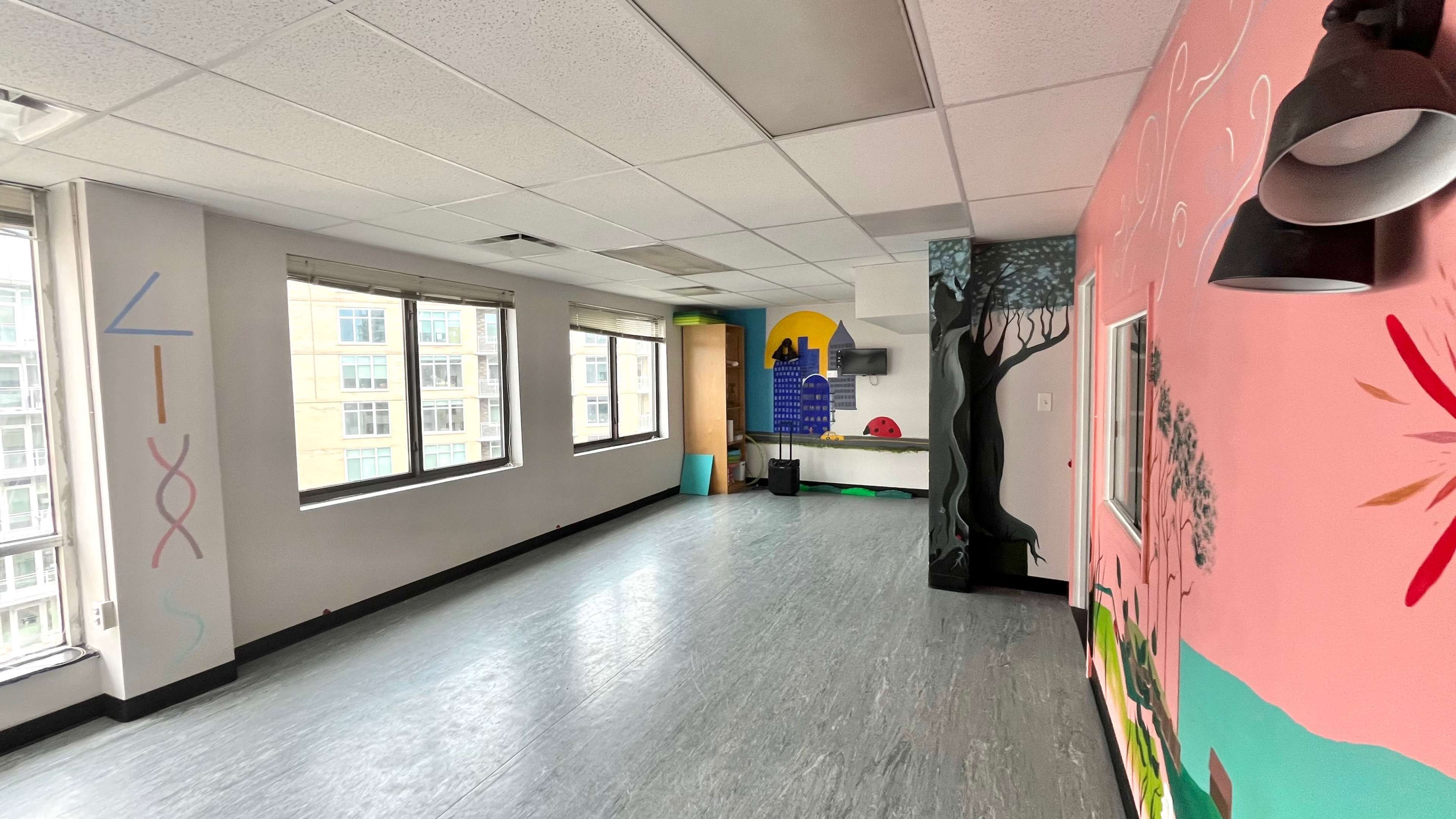 A brightly colored room with large windows, featuring a mural on one wall and a partially visible kitchenette area.