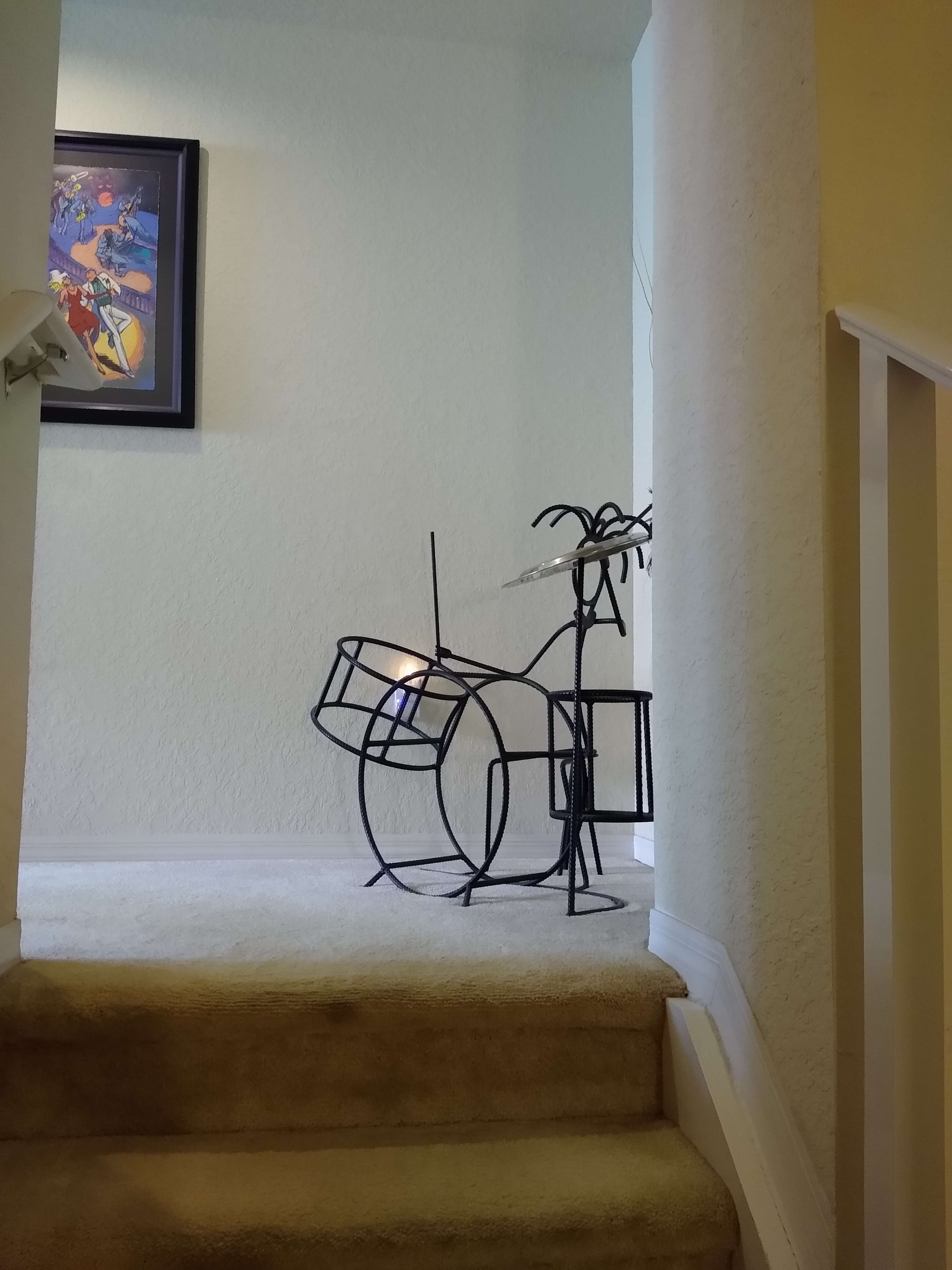 A metal sculpture of a drum set and a plant stands on a carpeted staircase near a wall with a framed artwork.