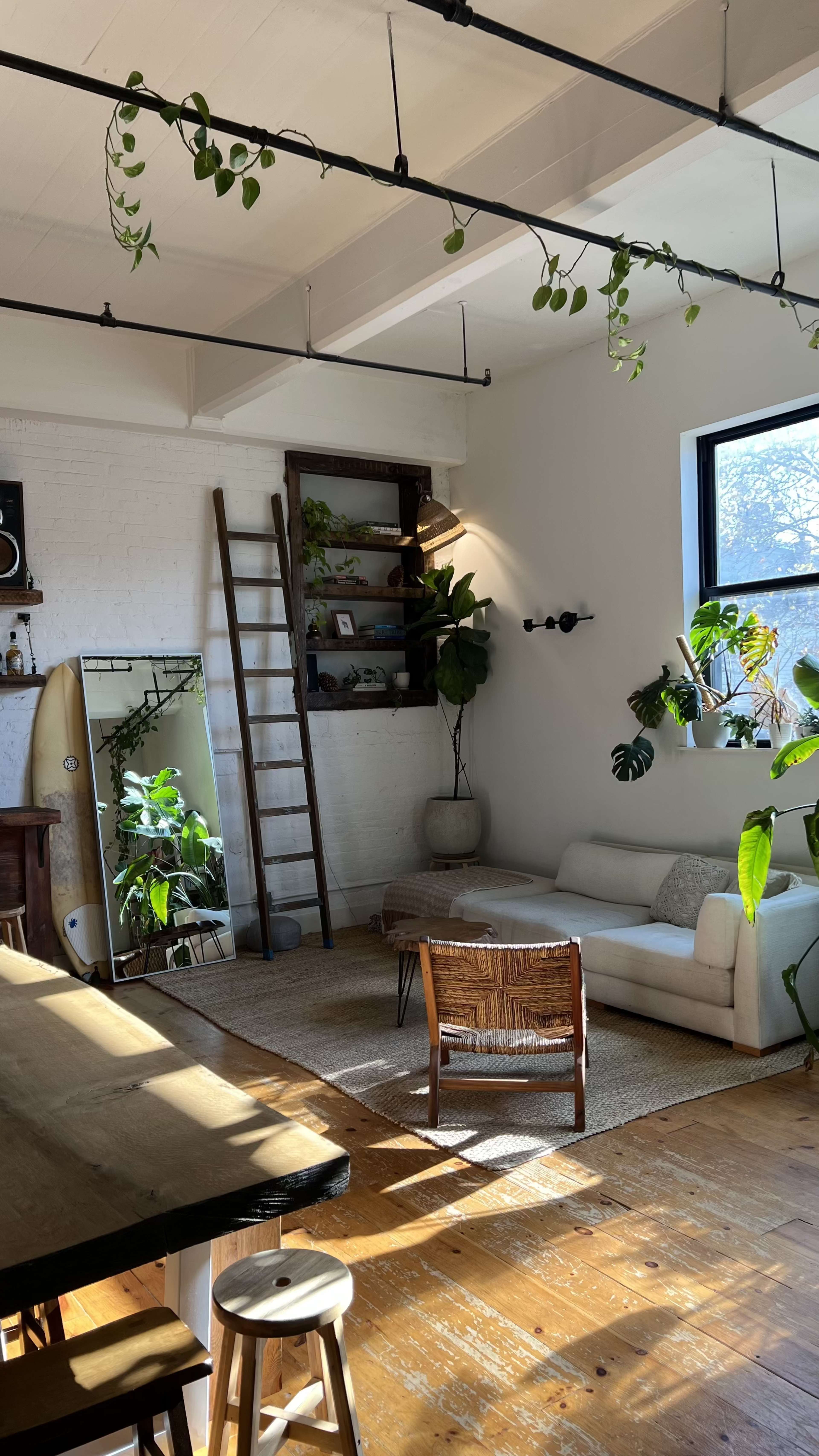 The image shows a bright living space with a white sofa, a rustic wooden dining table, plants placed strategically around the room, and a surfboard leaning against a wall.
