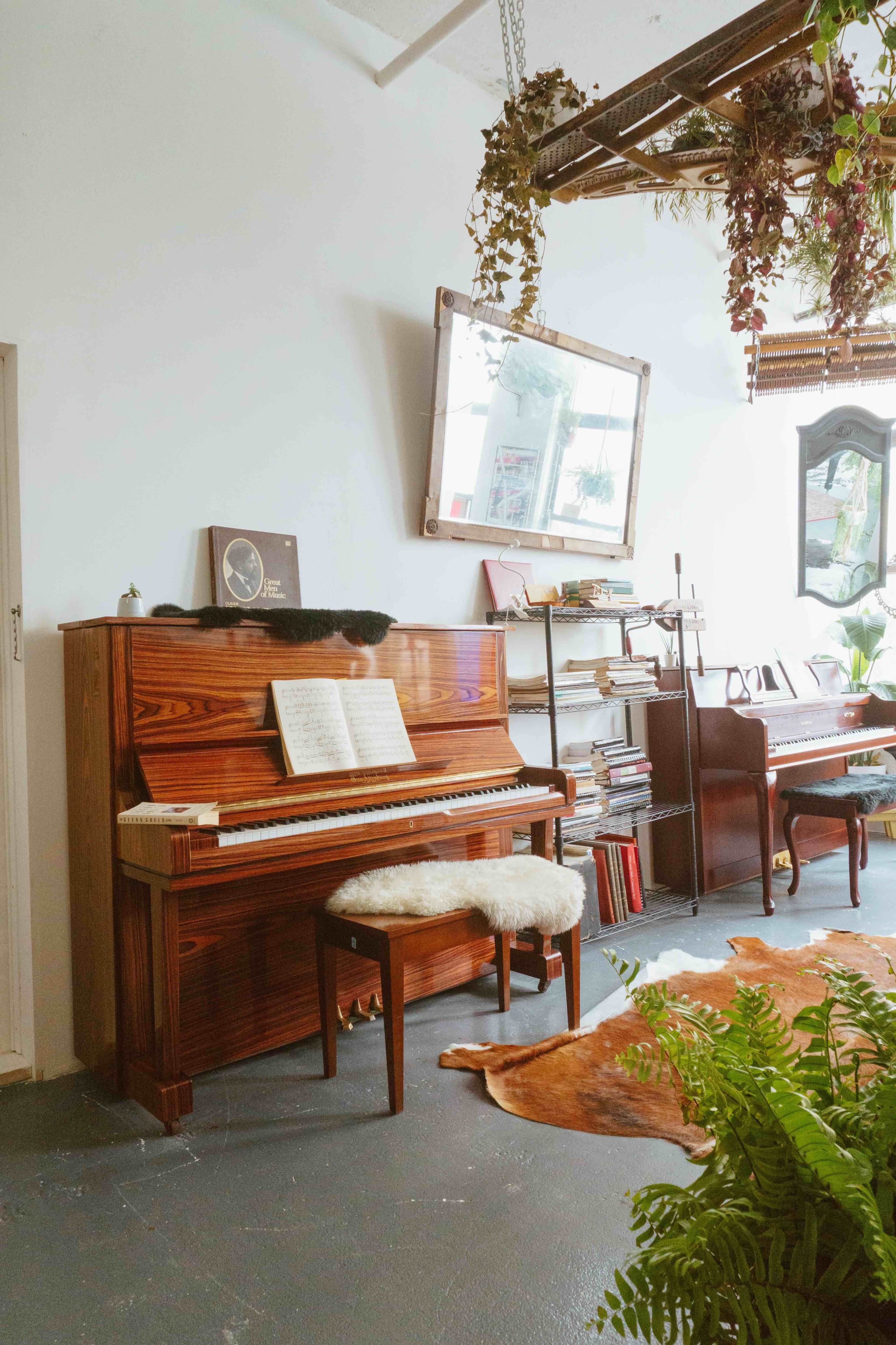 A wooden piano with sheet music sits beside bookshelves and a mirror in a room decorated with plants and a cowhide rug.