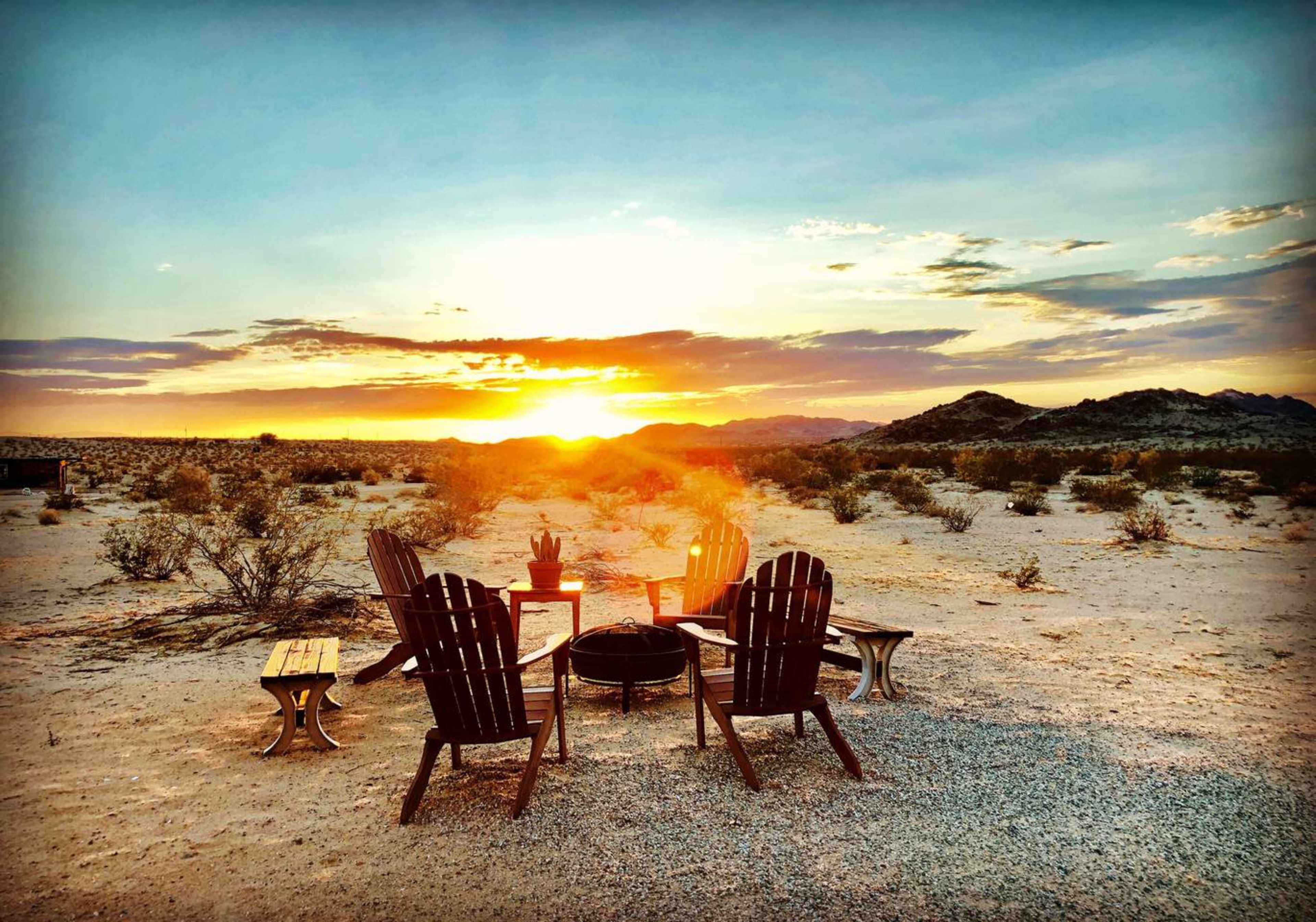A circle of four wooden chairs surrounds a fire pit in a desert landscape at sunset.