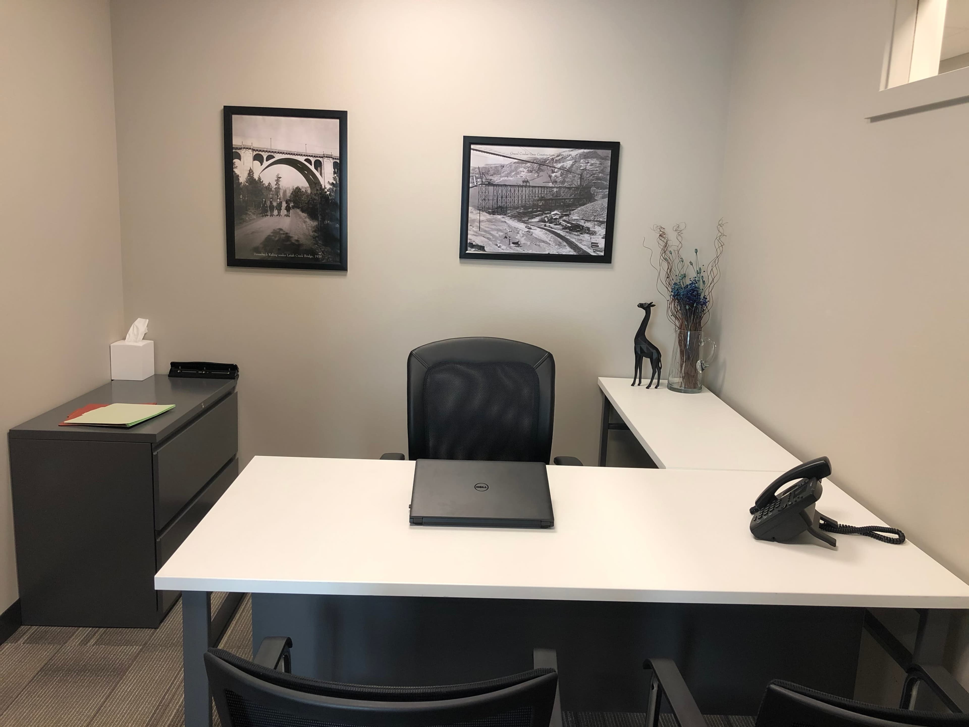 A modern office space features a desk with a laptop, a telephone, two framed black-and-white photographs on the walls, and a small decorative giraffe on the tabletop.