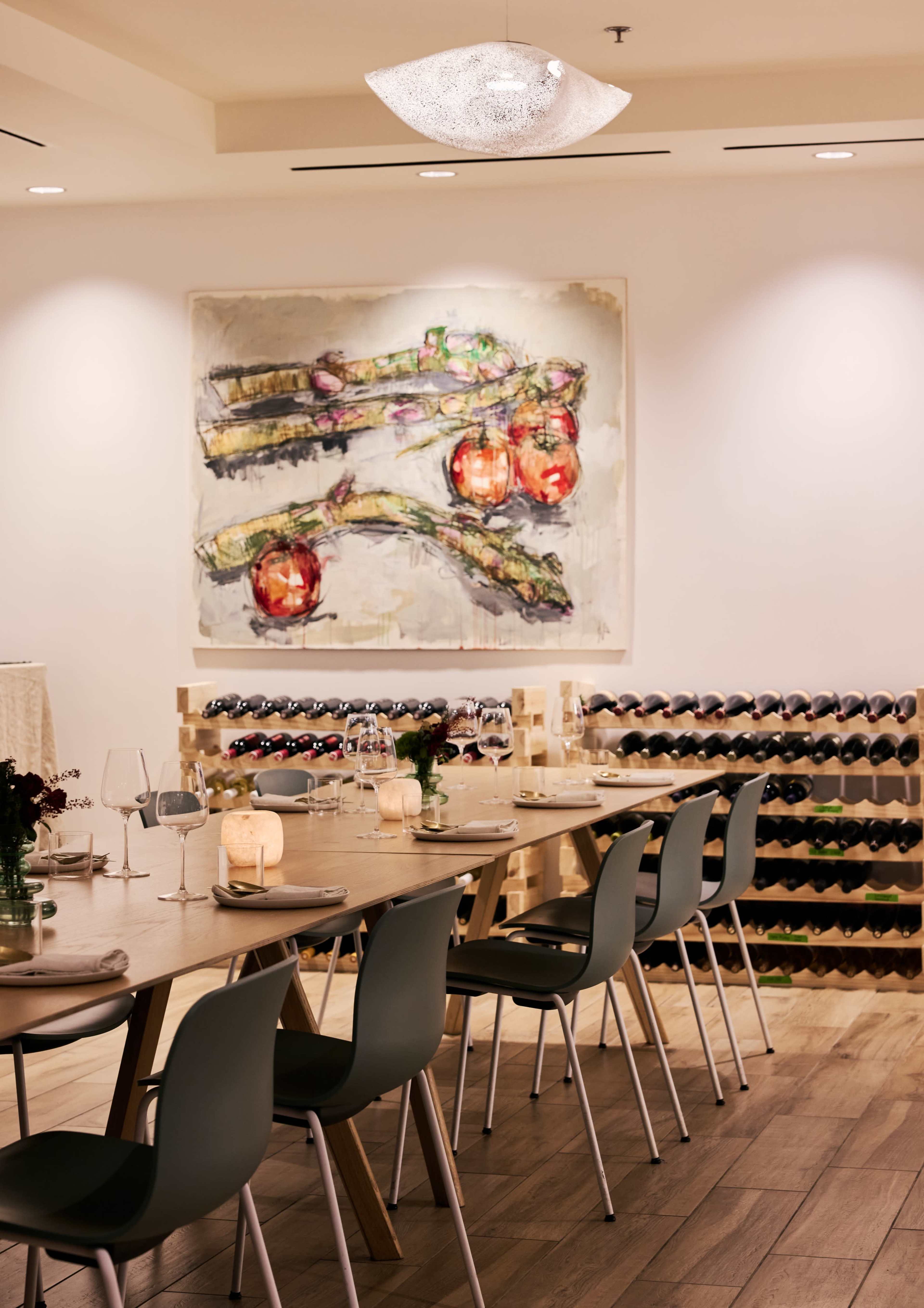 A wooden dining table is set with glassware and tableware, positioned near a wine rack and a large painting of apples on the wall.