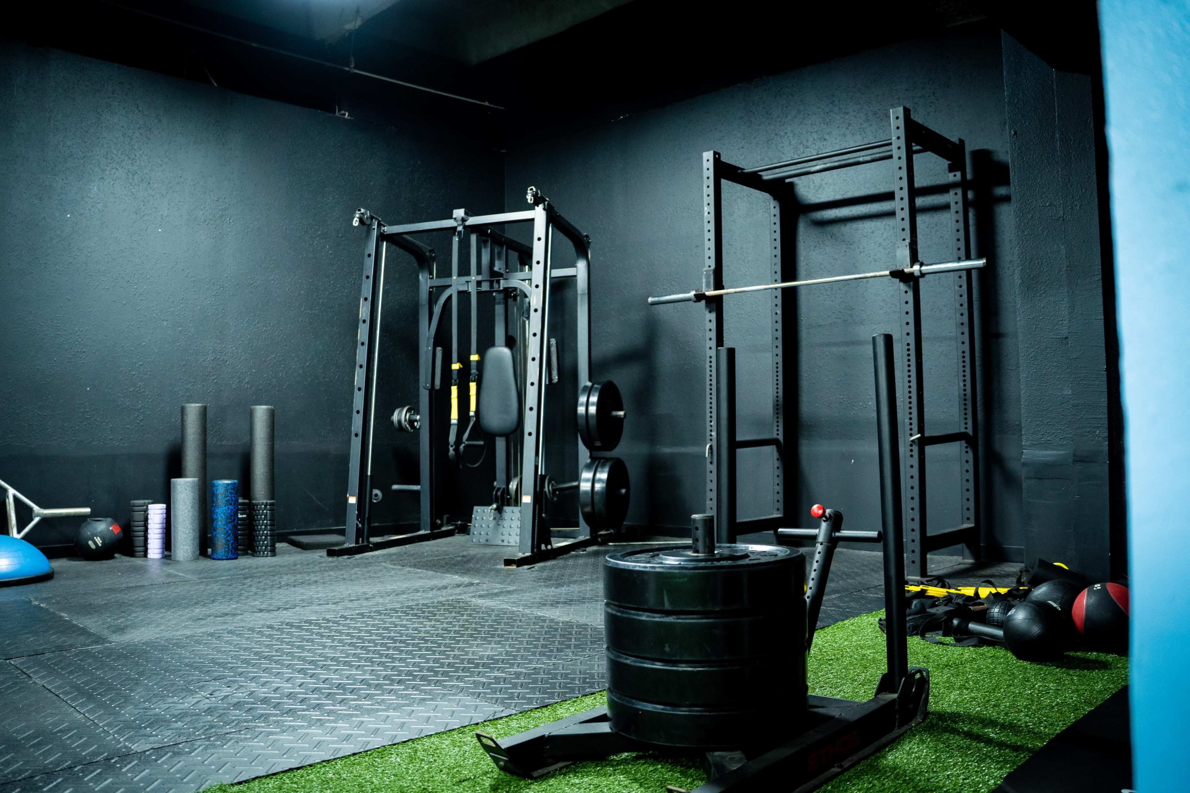 A dimly lit gym space featuring weightlifting equipment, including a power rack, a barbell on a squat rack, and various fitness accessories on a rubber floor.