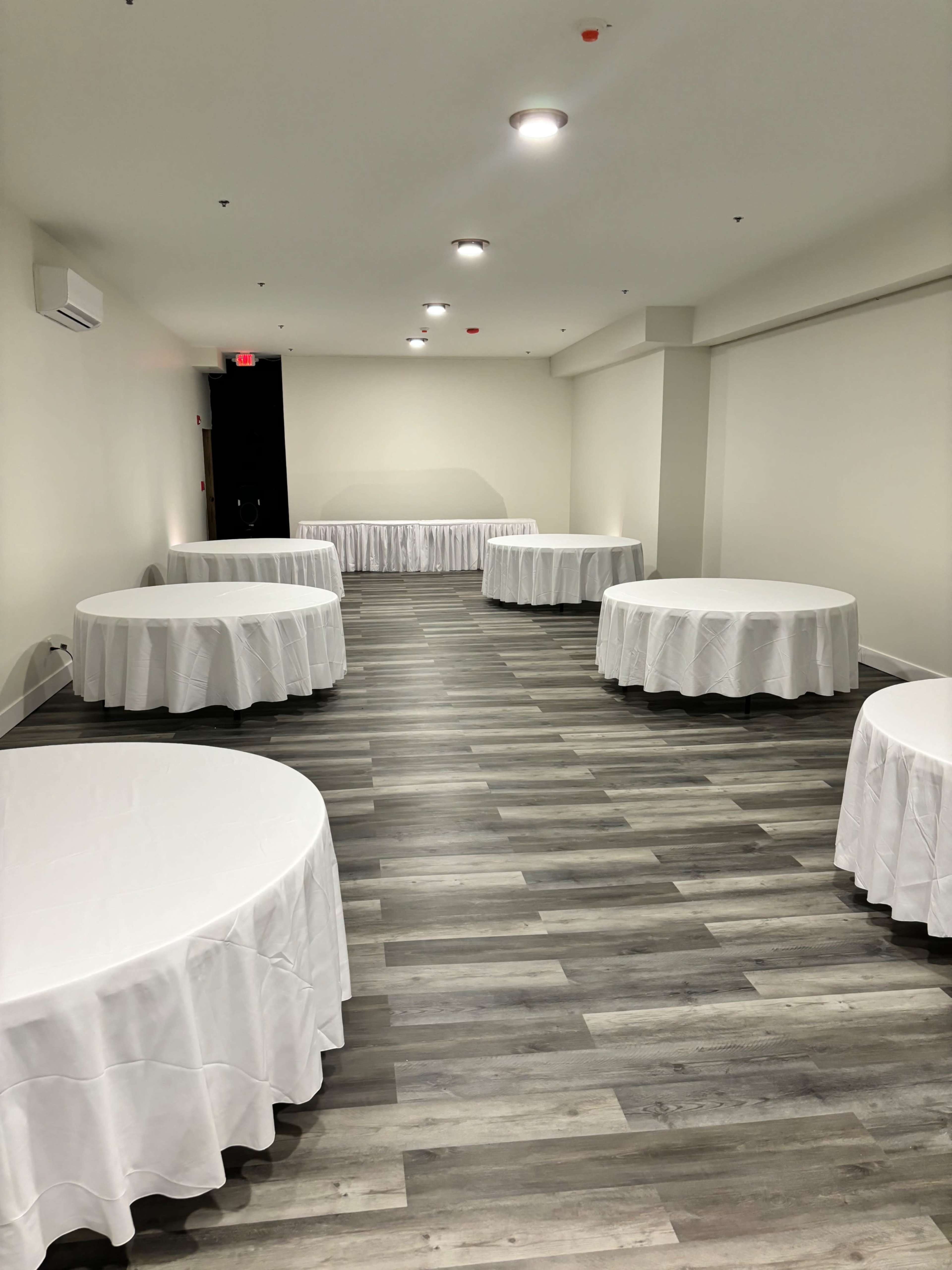 A spacious room features several round tables covered with white tablecloths, arranged around a polished wooden floor.
