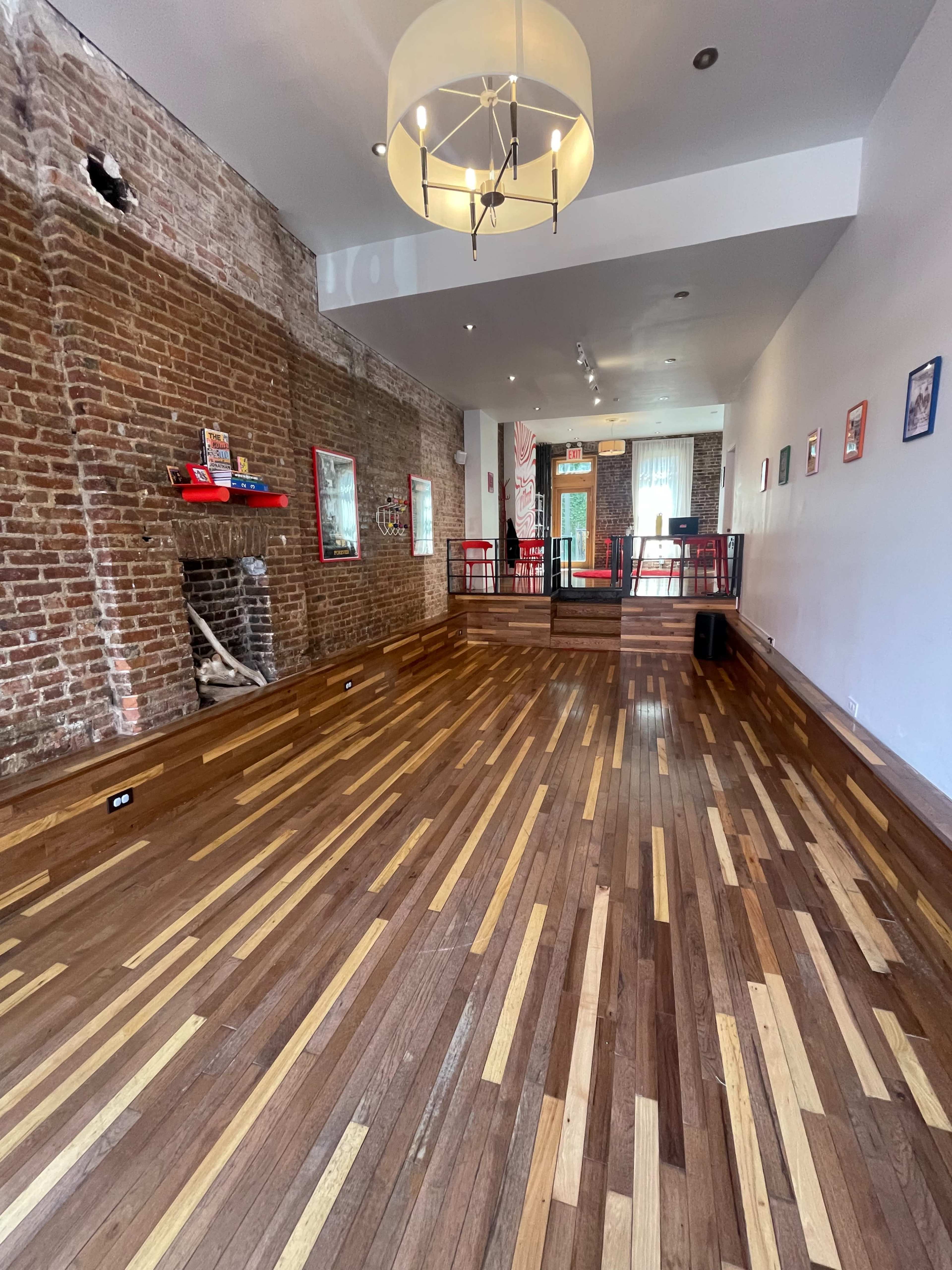 The image shows a spacious, empty room with wooden flooring and exposed brick walls, featuring a chandelier and a few framed pictures on the walls.