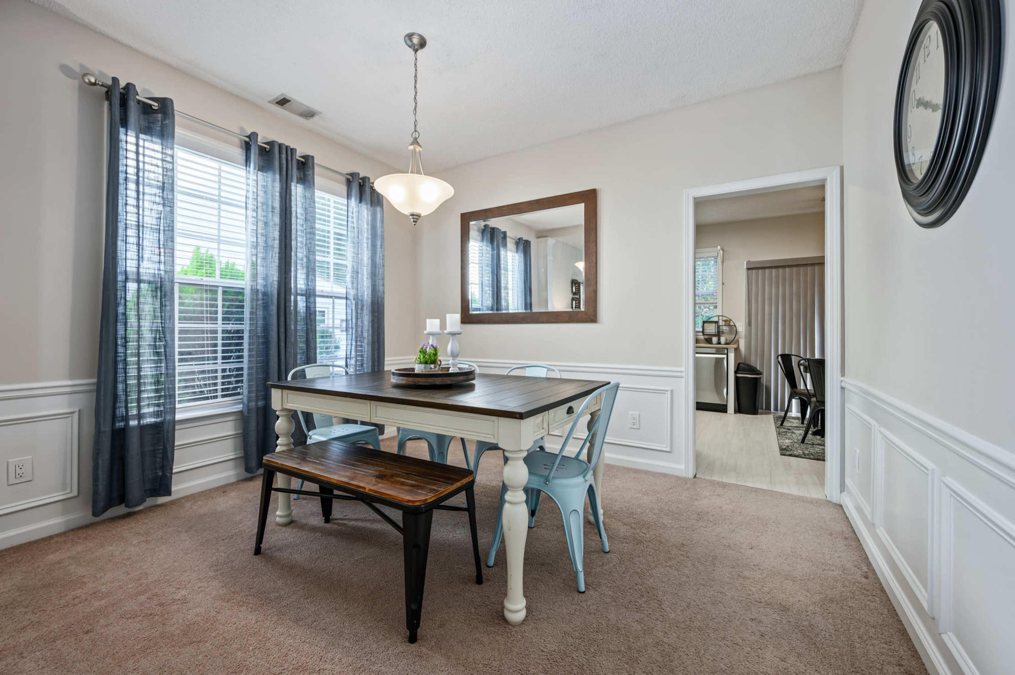 A dining area features a wooden table with a bench and chairs, alongside large windows dressed with dark curtains.