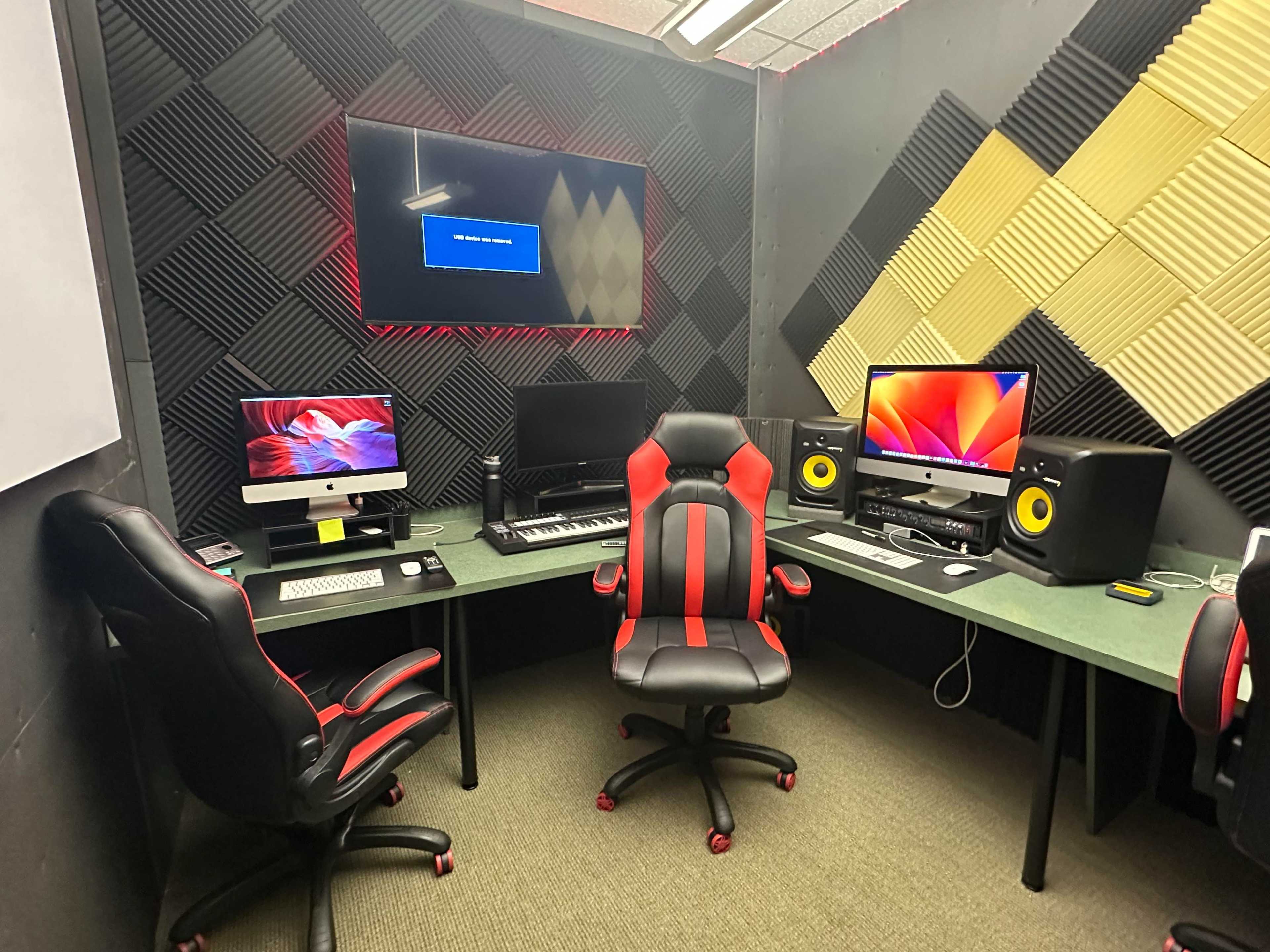 A music production studio with two computers, a keyboard, and speakers arranged around a green desk, all set against a soundproofed wall.