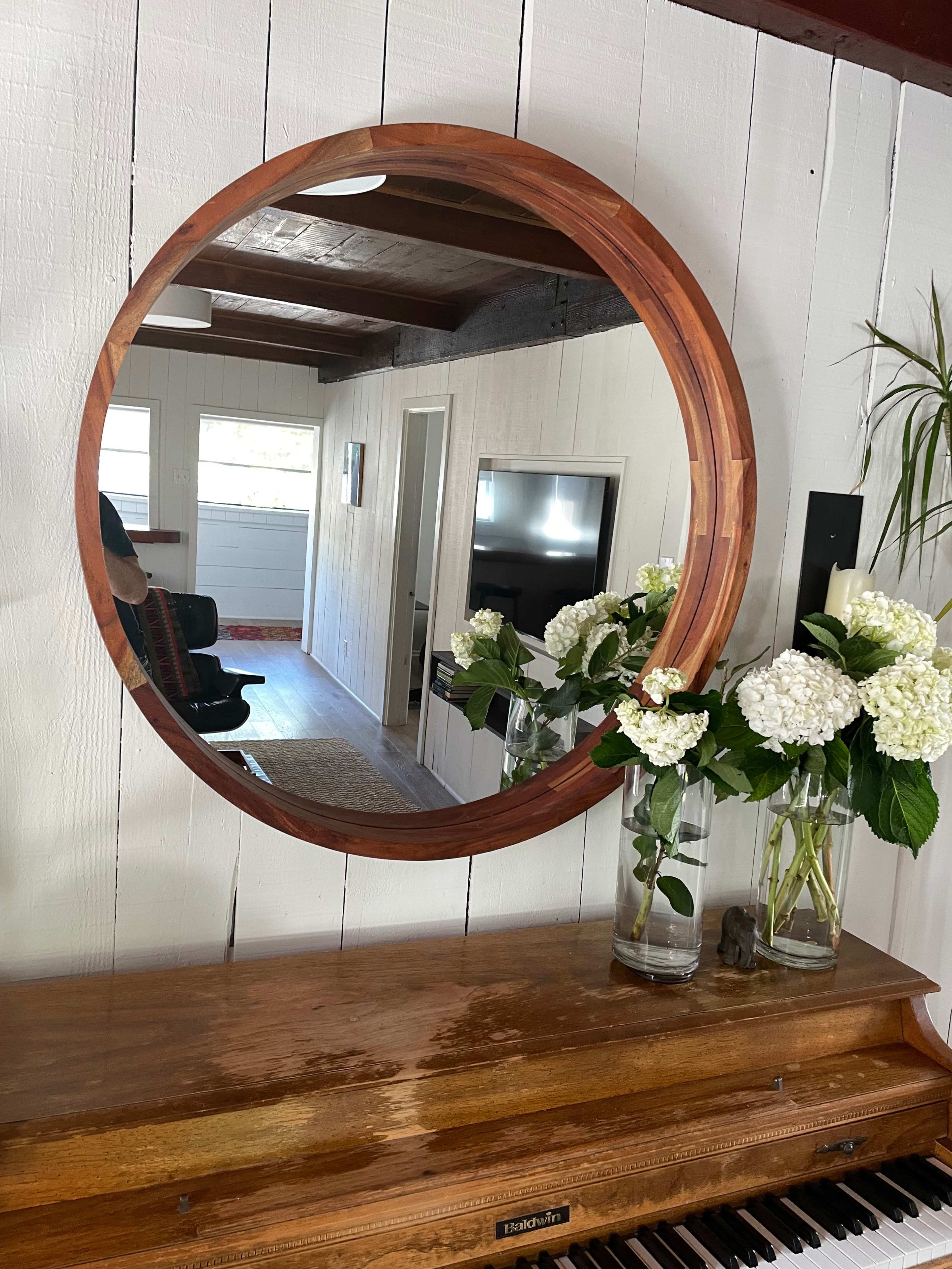 A round wooden mirror reflects a room with a piano, greenery, and light-colored walls.