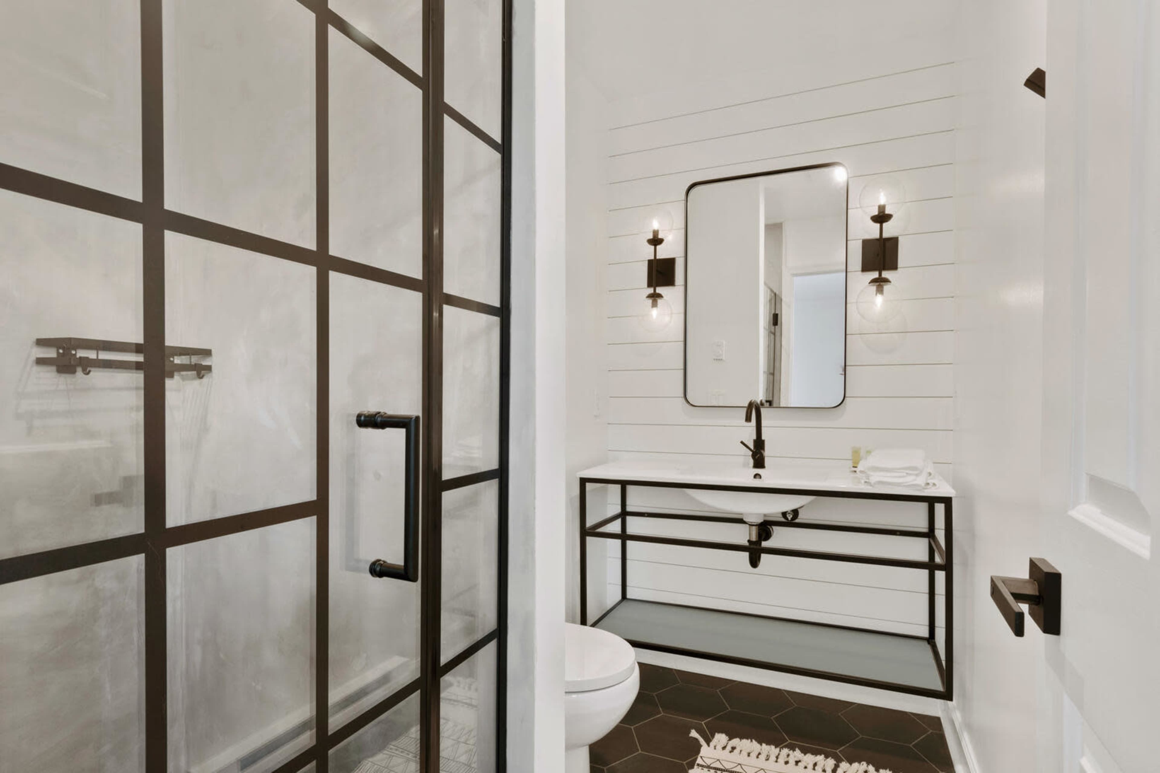 A modern bathroom with a black-framed glass shower, a white sink on a metal vanity, and a large mirror above it.