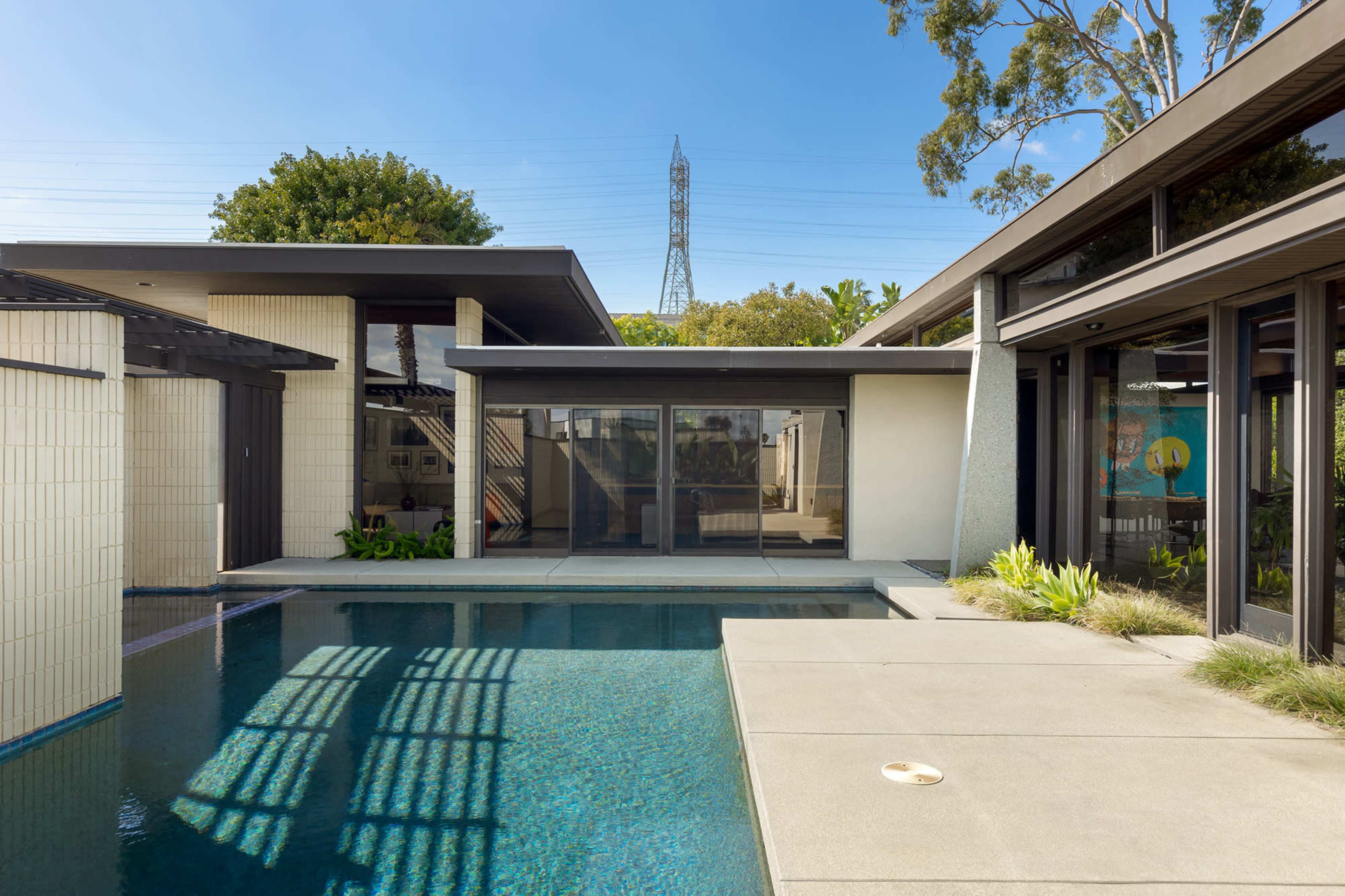 A modern house with large glass windows surrounds a rectangular swimming pool, featuring a patio area and greenery.