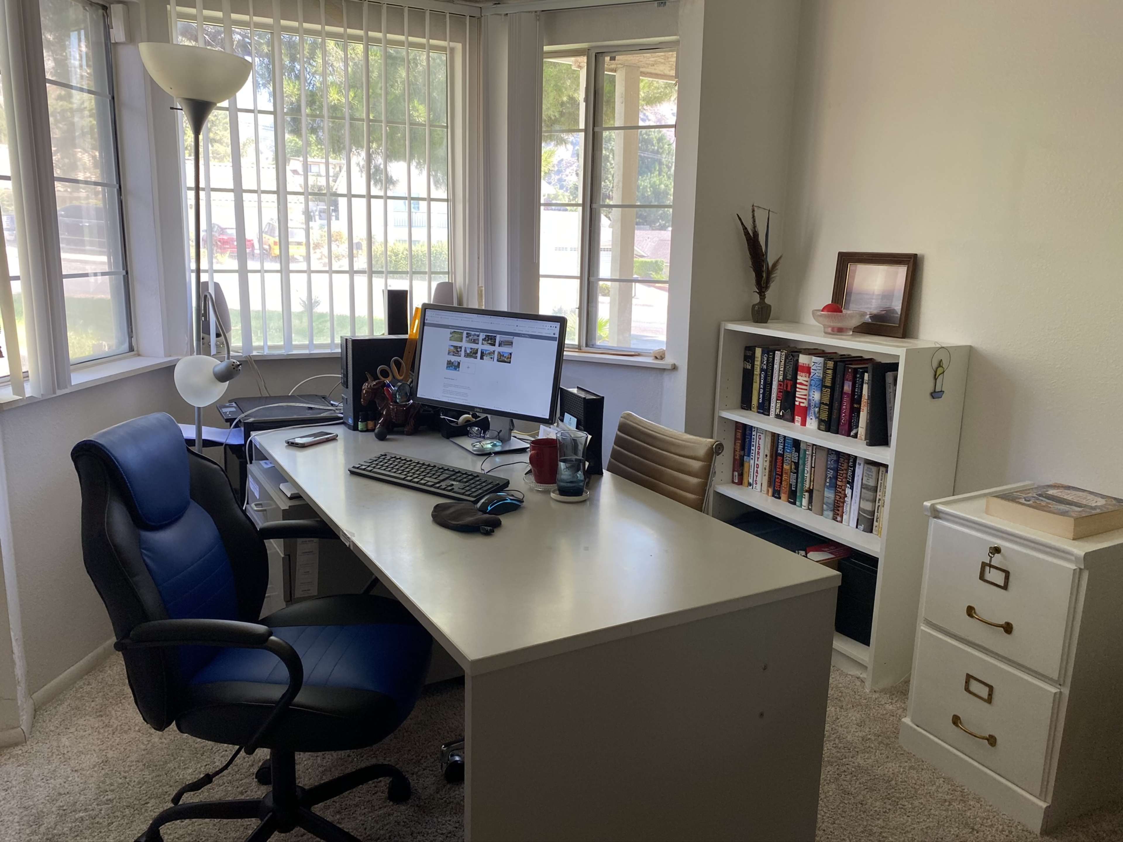 A home office features a desk with a computer, a chair, and a bookshelf against the wall, illuminated by natural light from nearby windows.