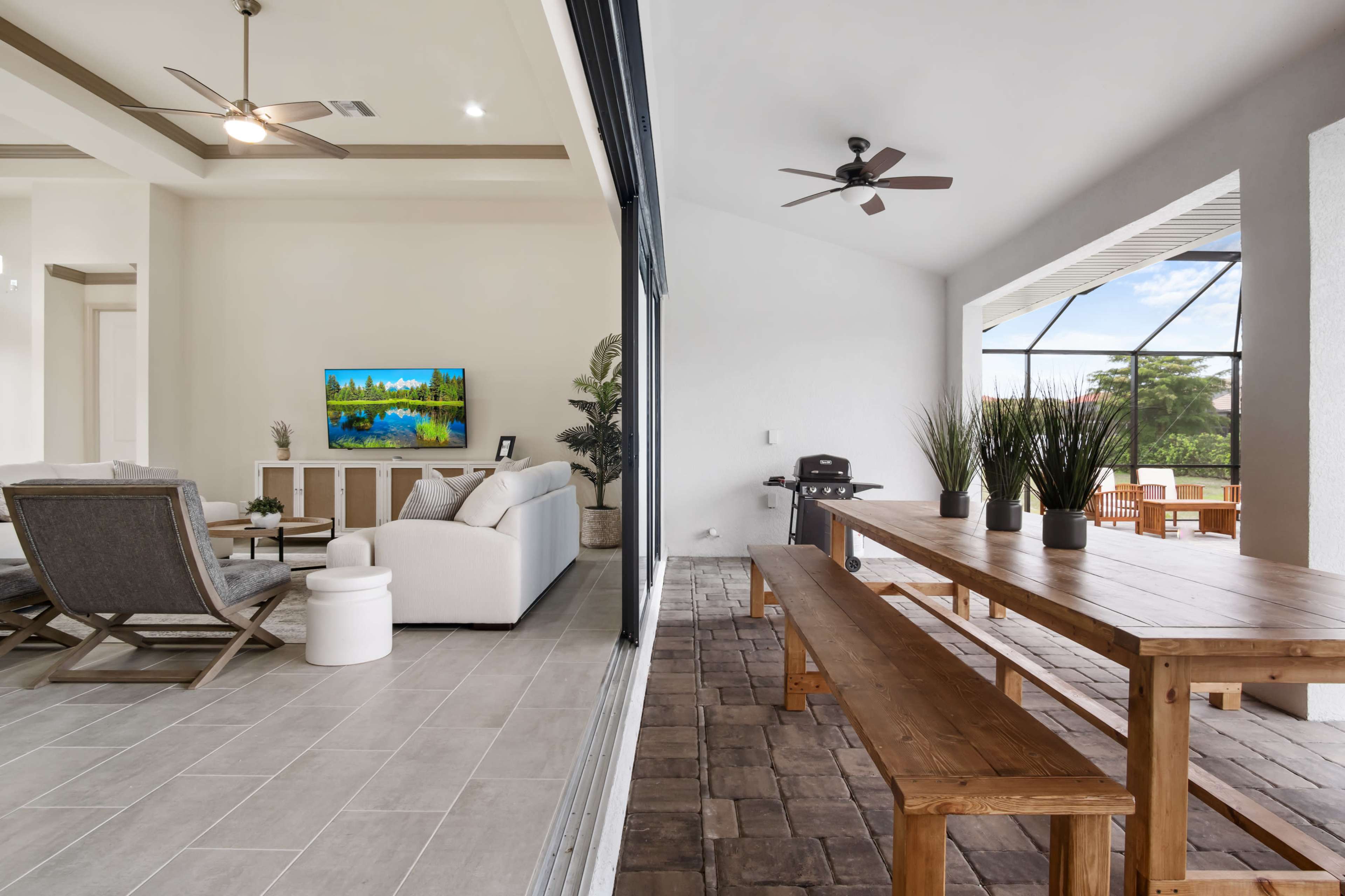 A large open space featuring a living area with a TV and seating, adjacent to a patio with a long wooden table and an outdoor view.