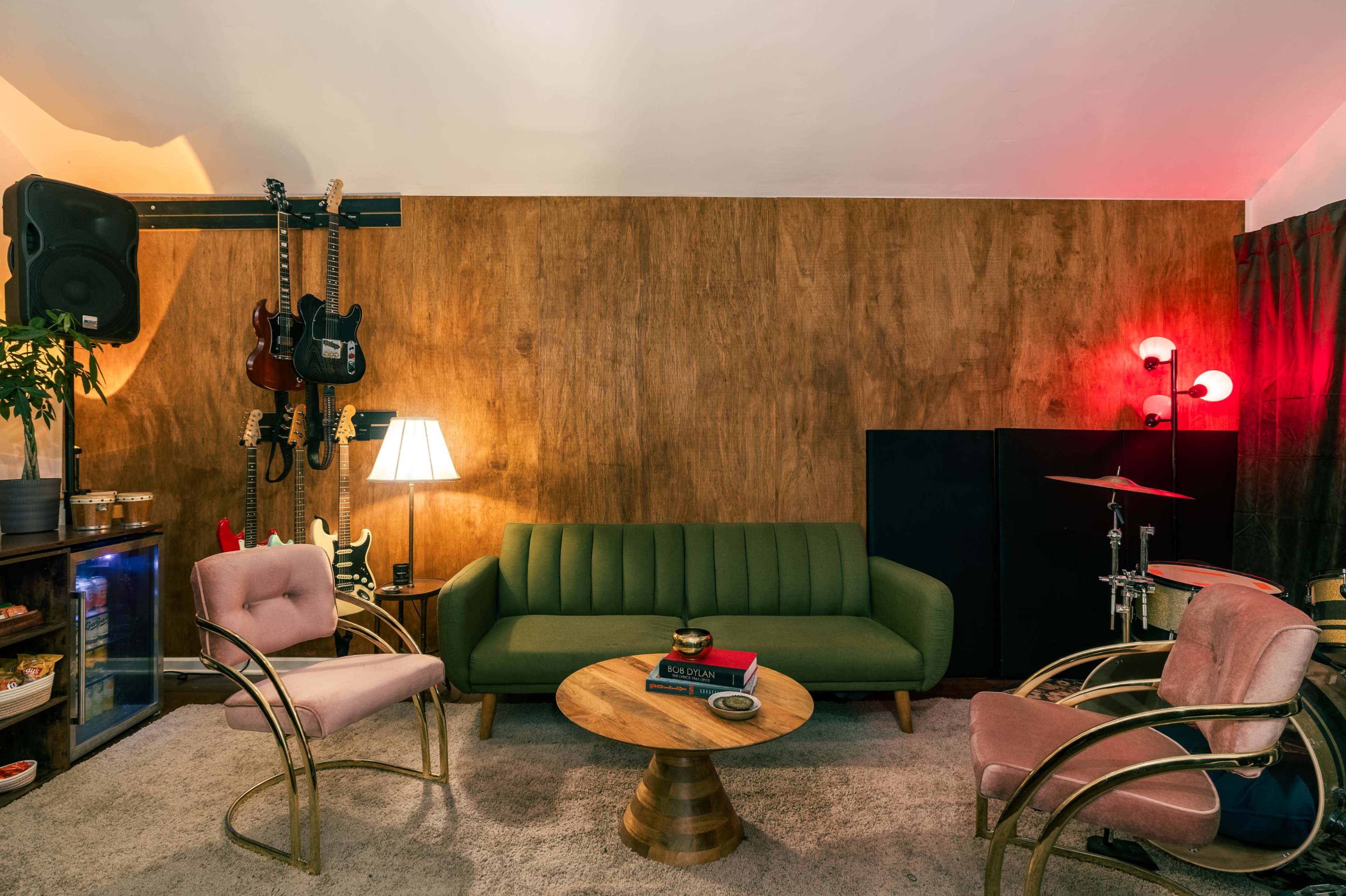 The image shows a cozy music-themed living room with a green sofa, two pink chairs, a wooden wall, and musical instruments displayed on the wall.