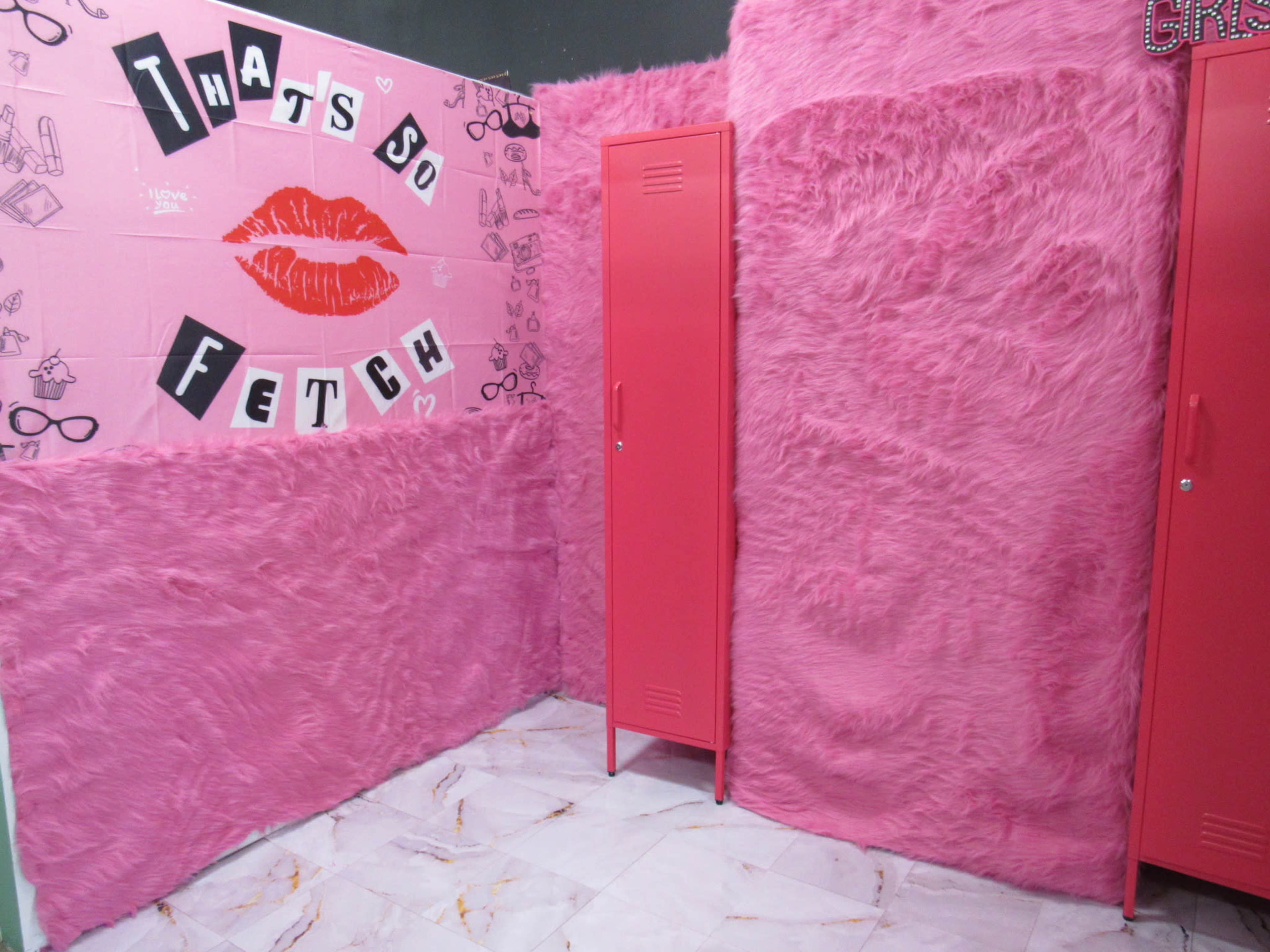 A brightly colored room with pink furry wall coverings and two pink lockers, featuring a wall that has a collage with the phrase "THAT'S SO FETCH."