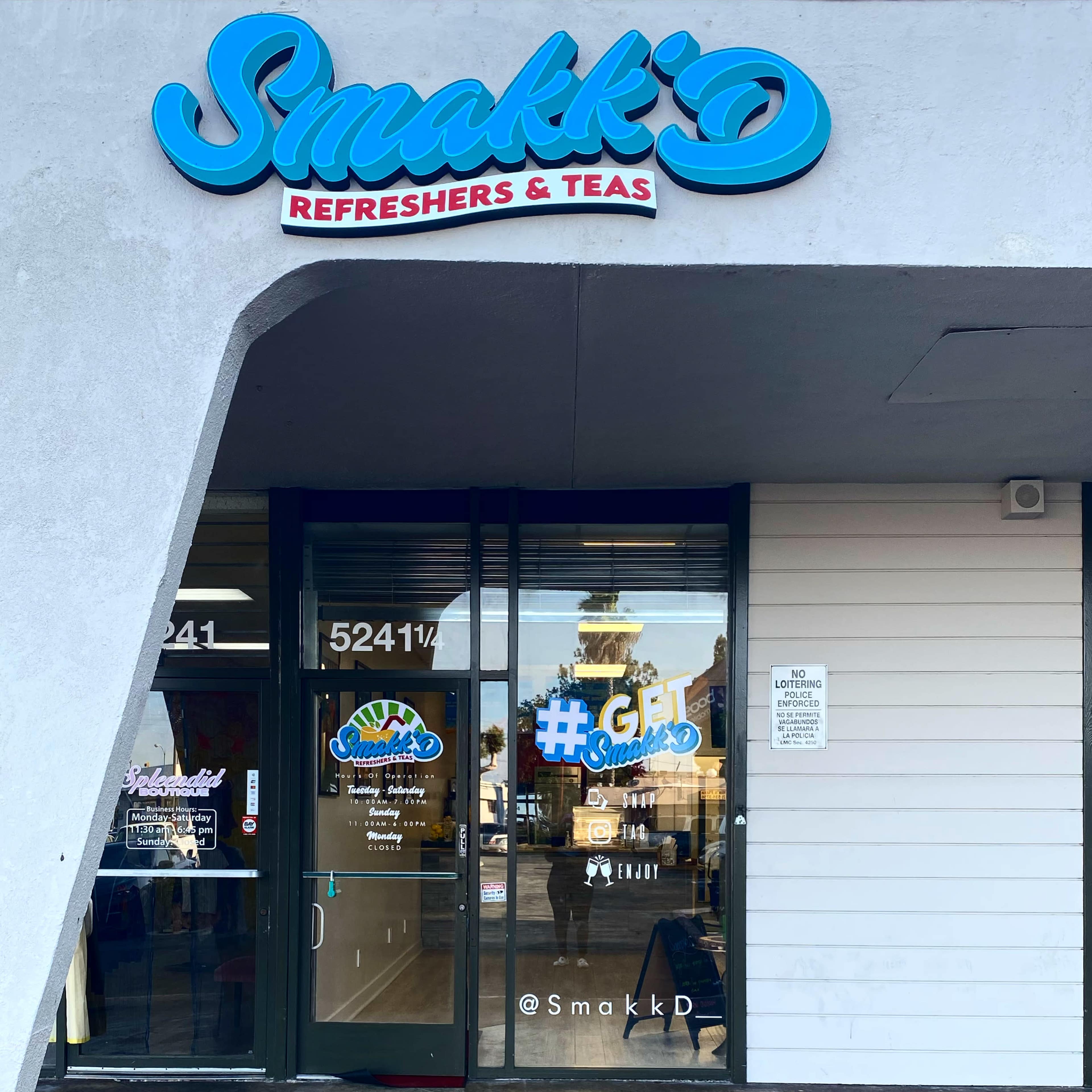 The entrance of a shop called "Smakk'D Refreshers & Teas," featuring a blue and white sign and a large glass door with visible interior displays.