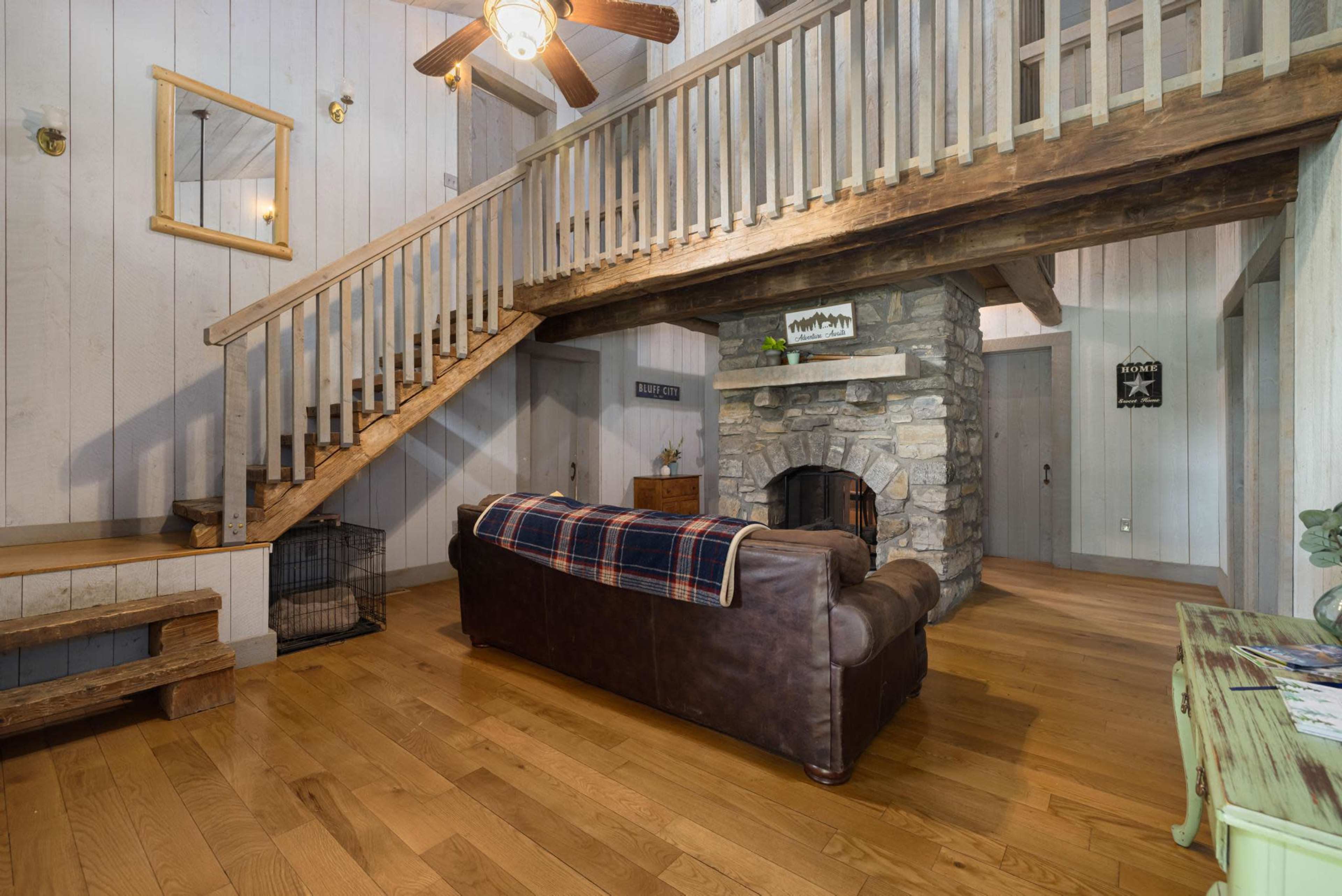 The image shows a wooden interior with a staircase, a stone fireplace, a brown leather couch, and natural light coming through a window.