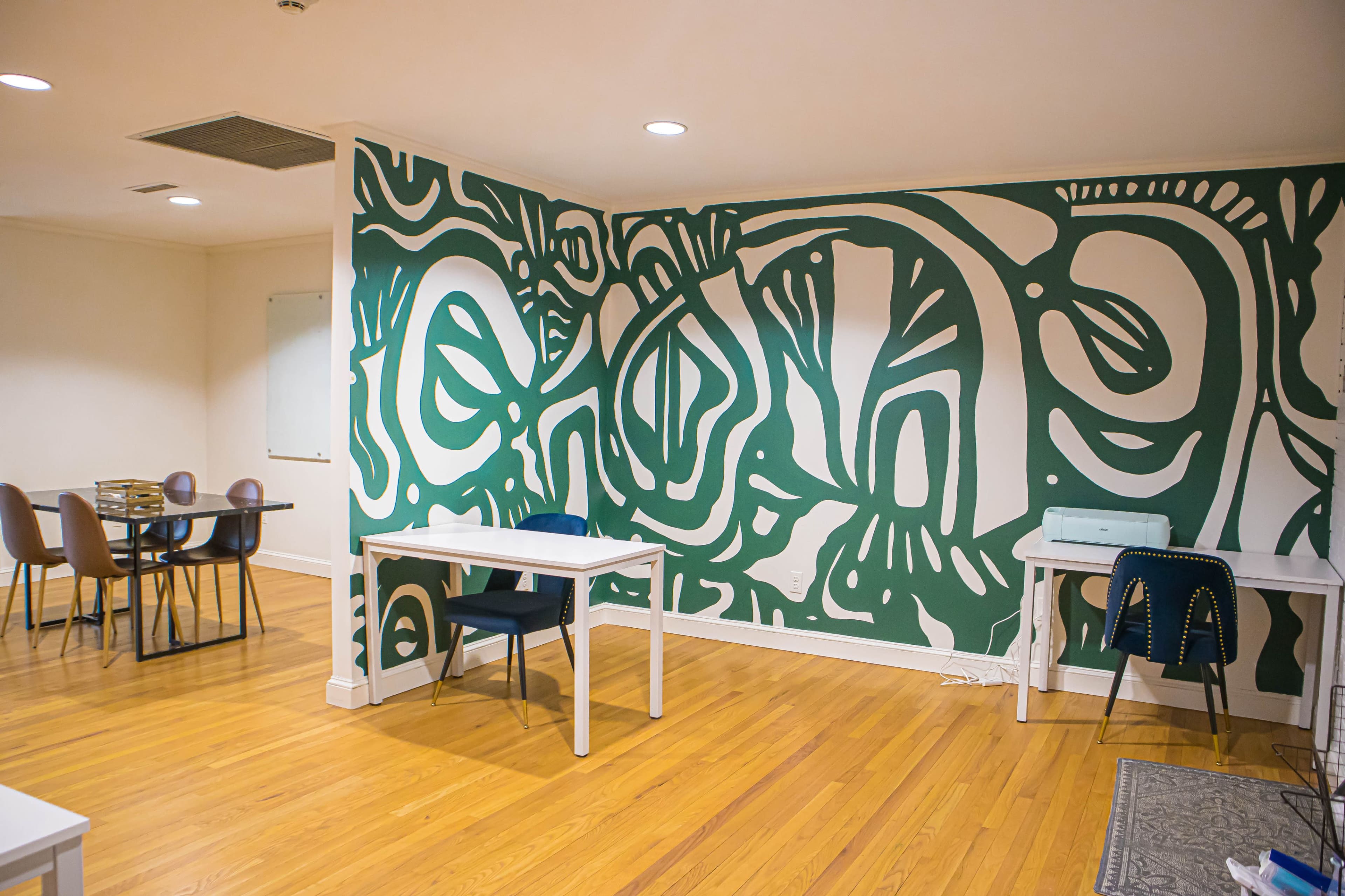 The image shows a room with a green abstract mural on one wall, featuring tables and chairs arranged for workspace.
