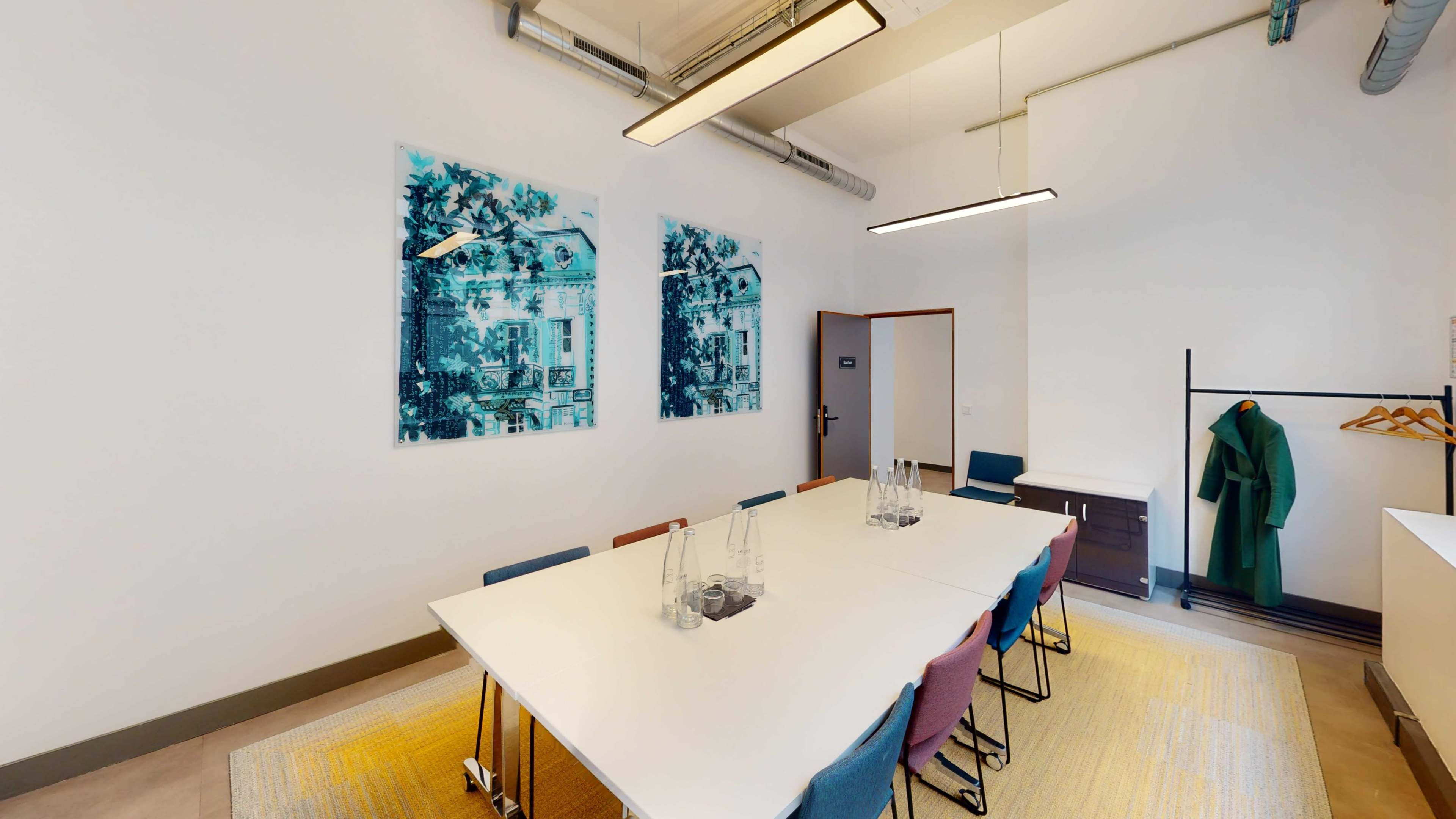 A modern meeting room features a long, white table surrounded by colored chairs, with artwork on the walls and a coat rack in the corner.