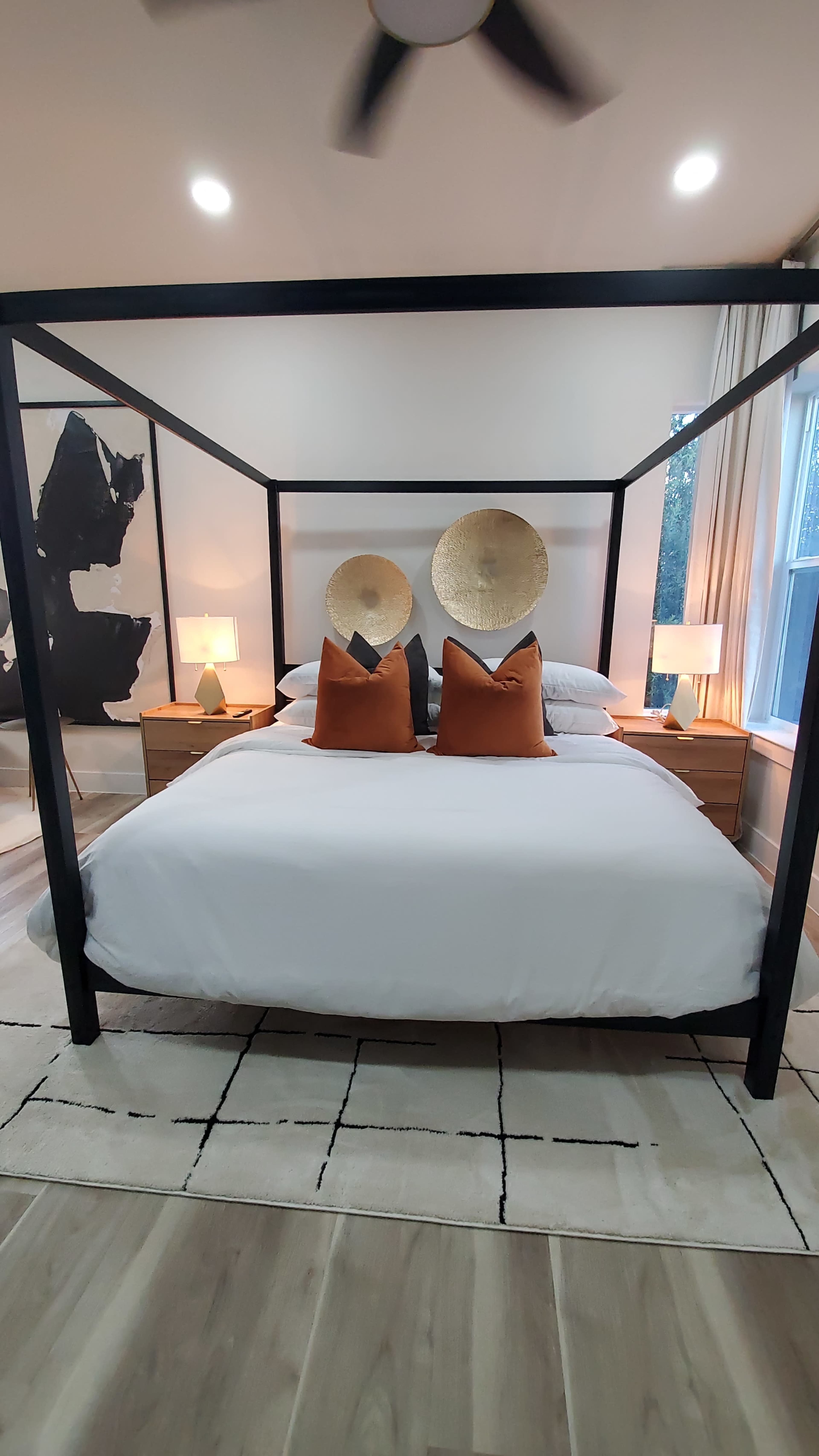 The image shows a modern bedroom with a four-poster bed, two decorative circular wall pieces above the headboard, and bedside tables with lamps on either side.
