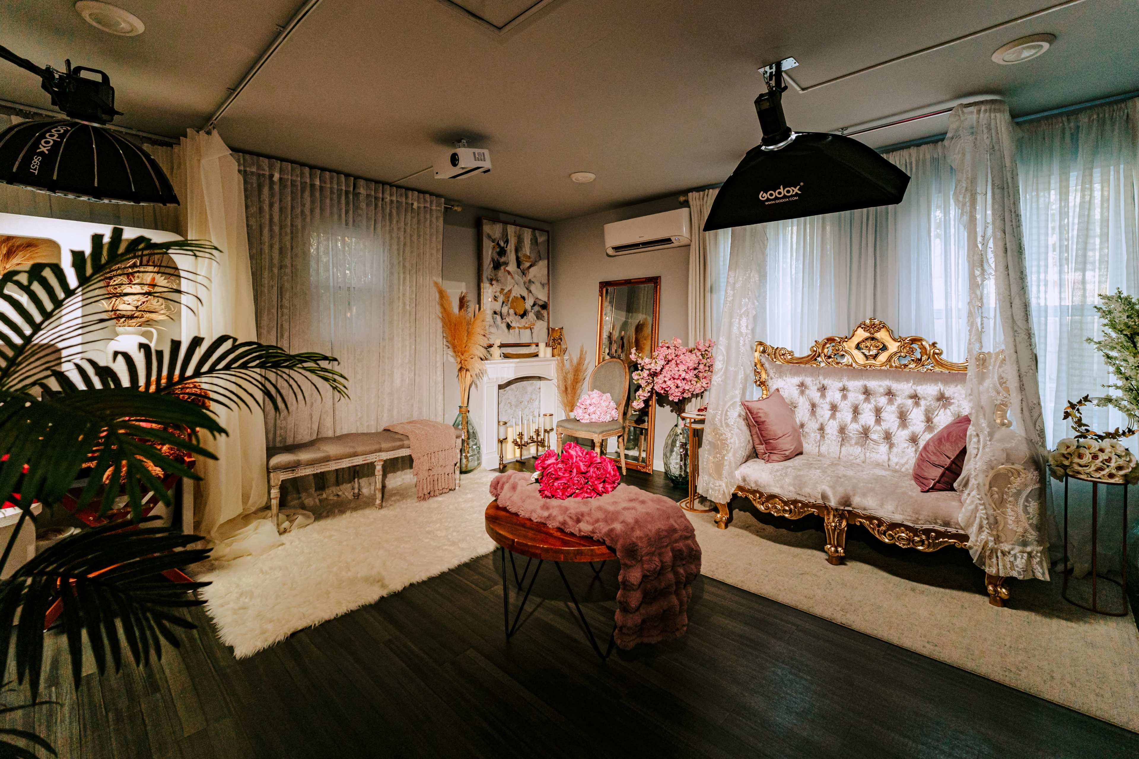 The image shows a stylish interior living space featuring a gold-accented sofa, a plush rug, and decorative plants, with soft lighting and draped curtains.