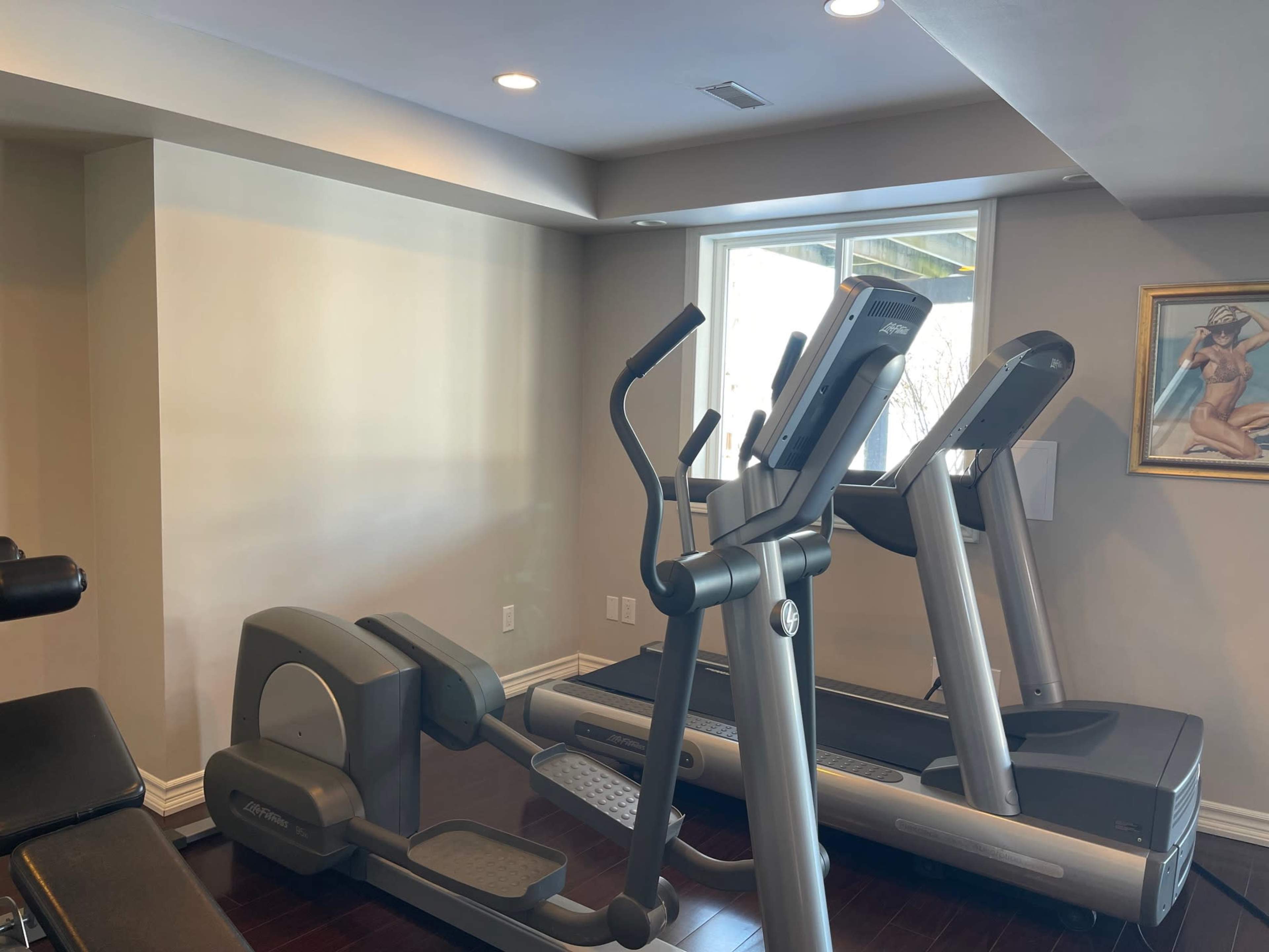 The image shows a home gym with a treadmill, an elliptical machine, and a weight bench, all set against a wall with a window and a framed artwork.
