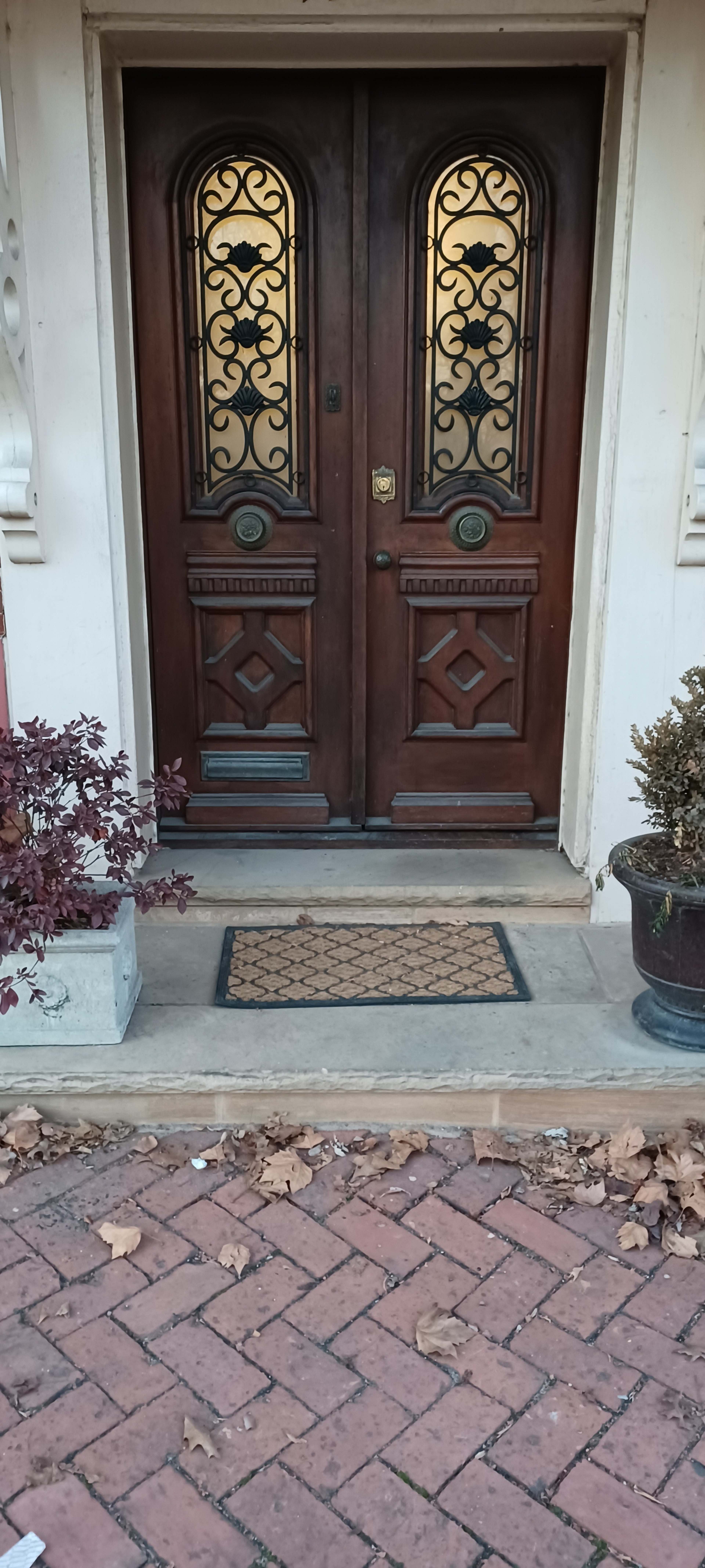 A pair of ornate wooden doors with decorative glass panels, flanked by potted plants and a welcome mat on a brick pathway.