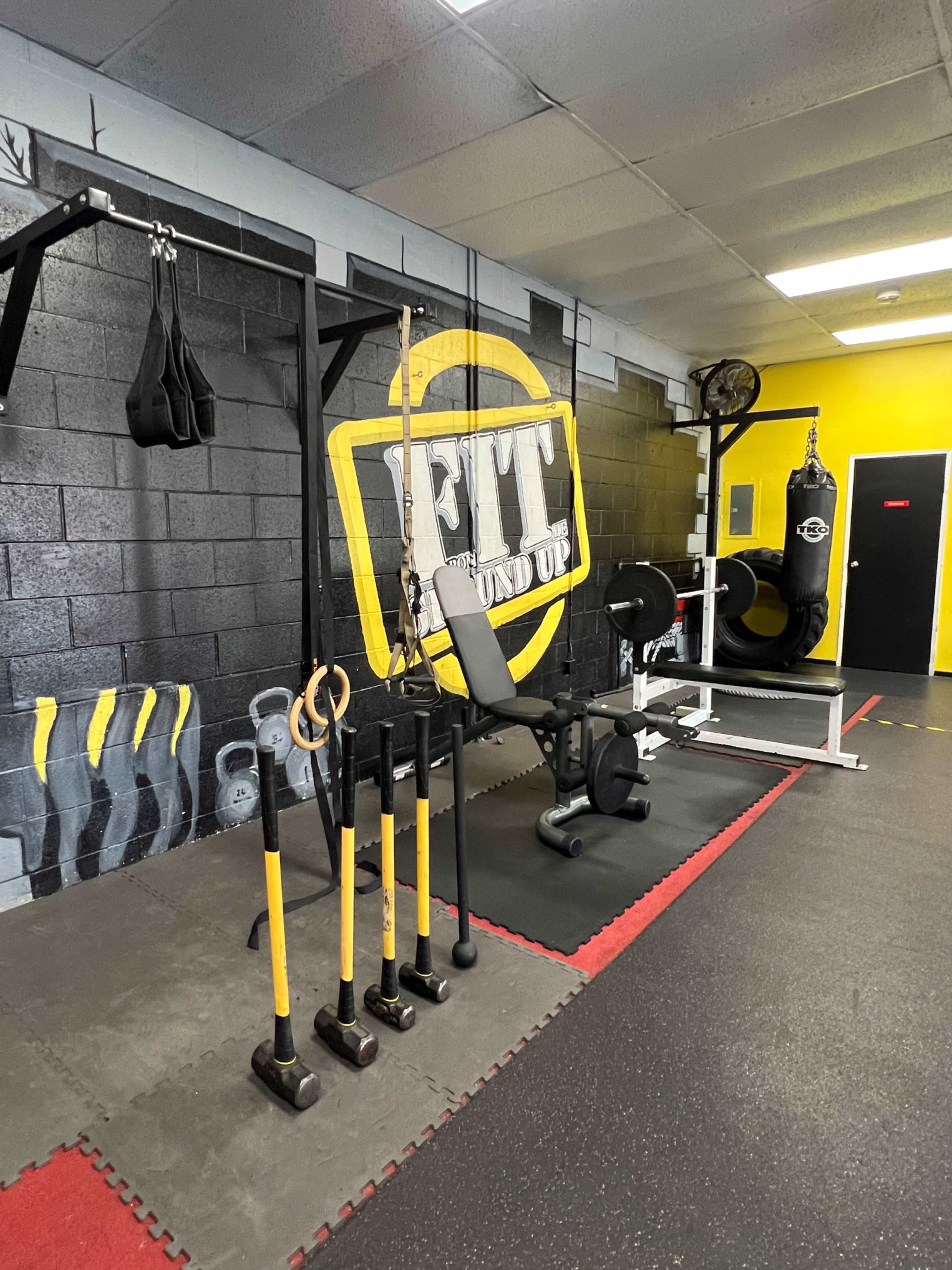 The image shows a gym interior featuring various workout equipment, including weightlifting benches, a punching bag, and exercise tools hanging on a wall with a prominent "FIT" logo.