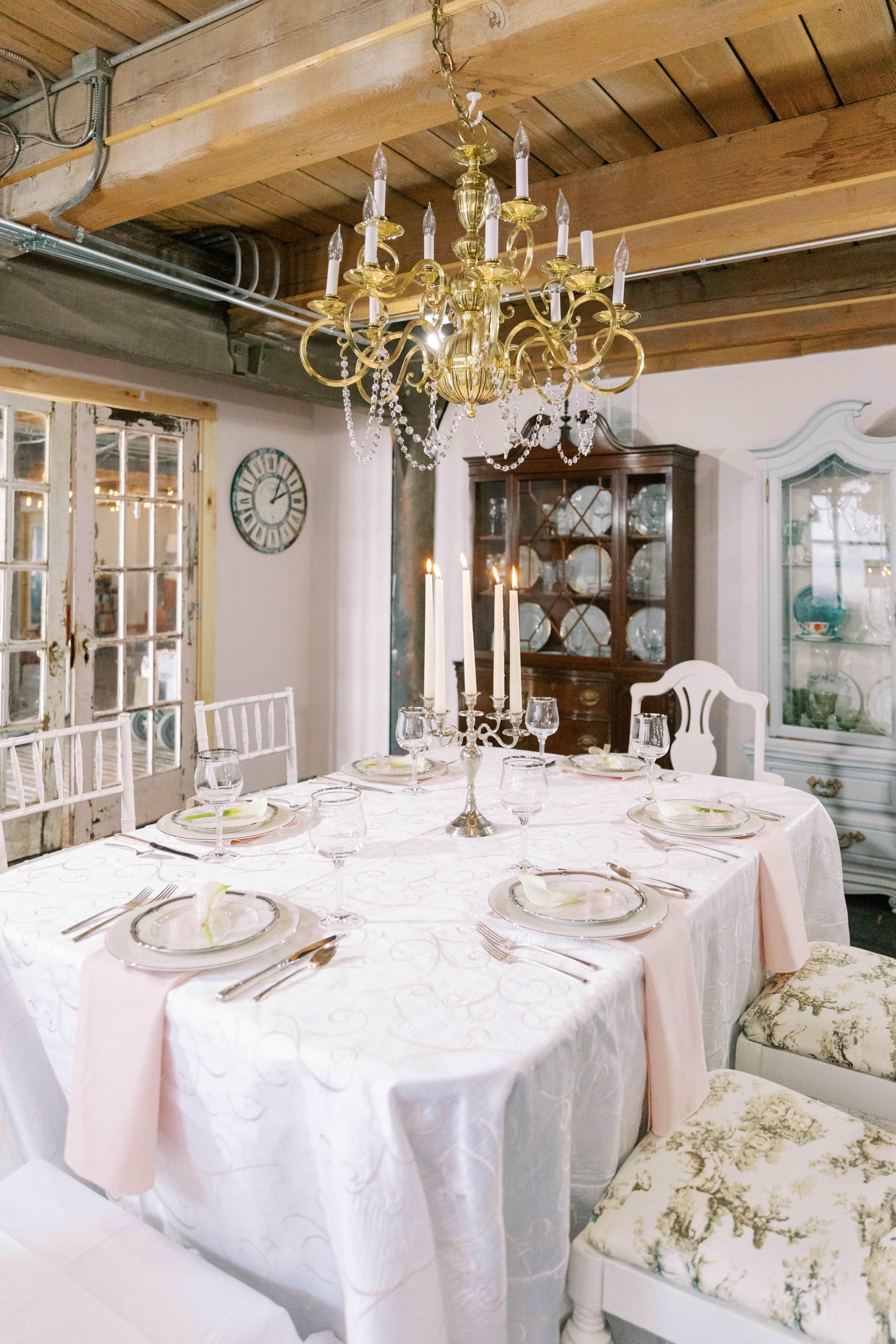 A dining table is set with elegant tableware and a chandelier hangs overhead in a well-lit room.